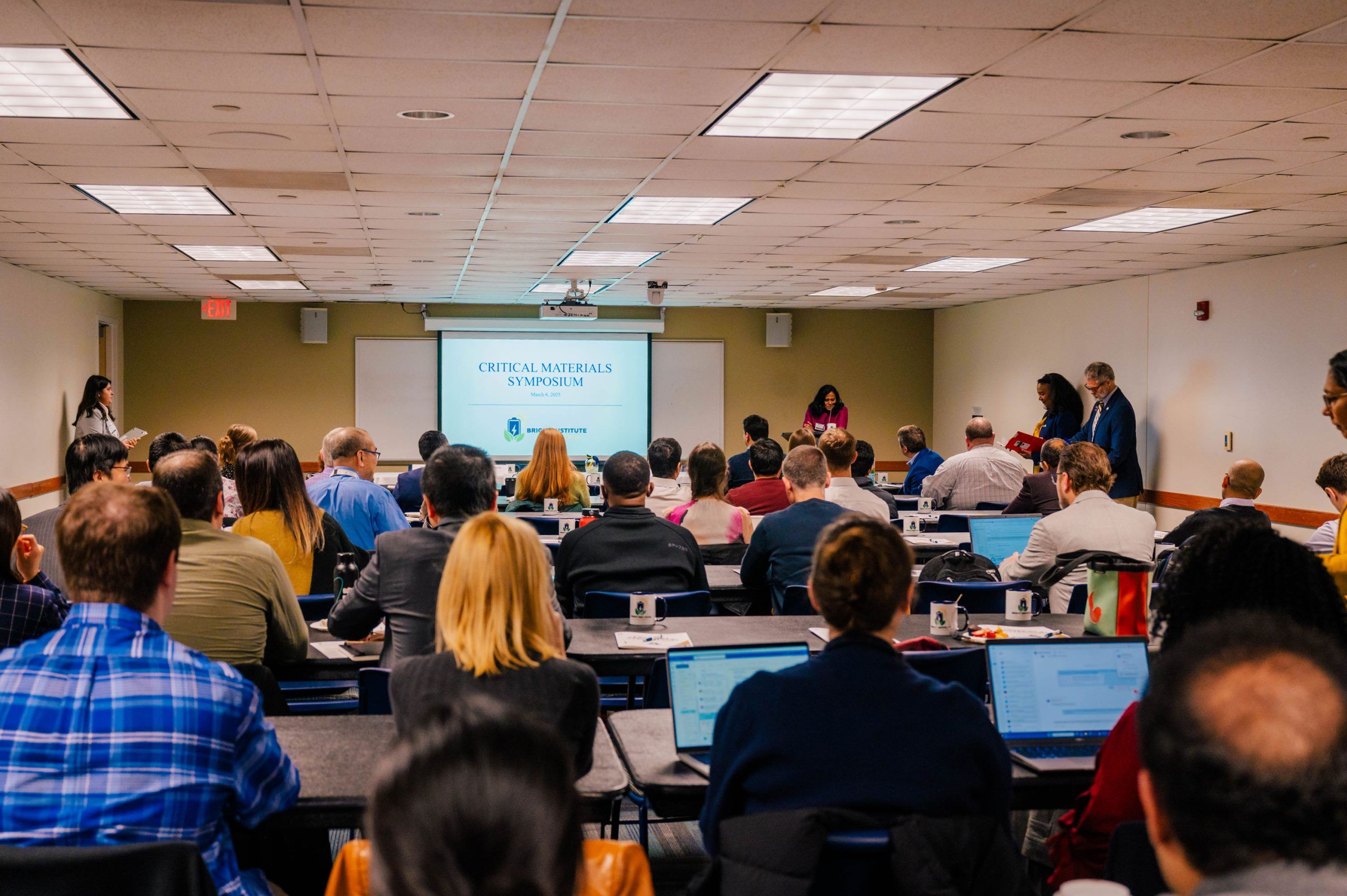 A conference room filled with attendees seated at tables, facing a projection screen at the front that displays the title “Critical Materials Symposium” along with the Bright Institute and UNC Greensboro logos. Laptops, notebooks, and coffee cups are visible on the tables. Several people stand near the front and side of the room, and the setting has a drop ceiling with fluorescent lighting and beige walls.