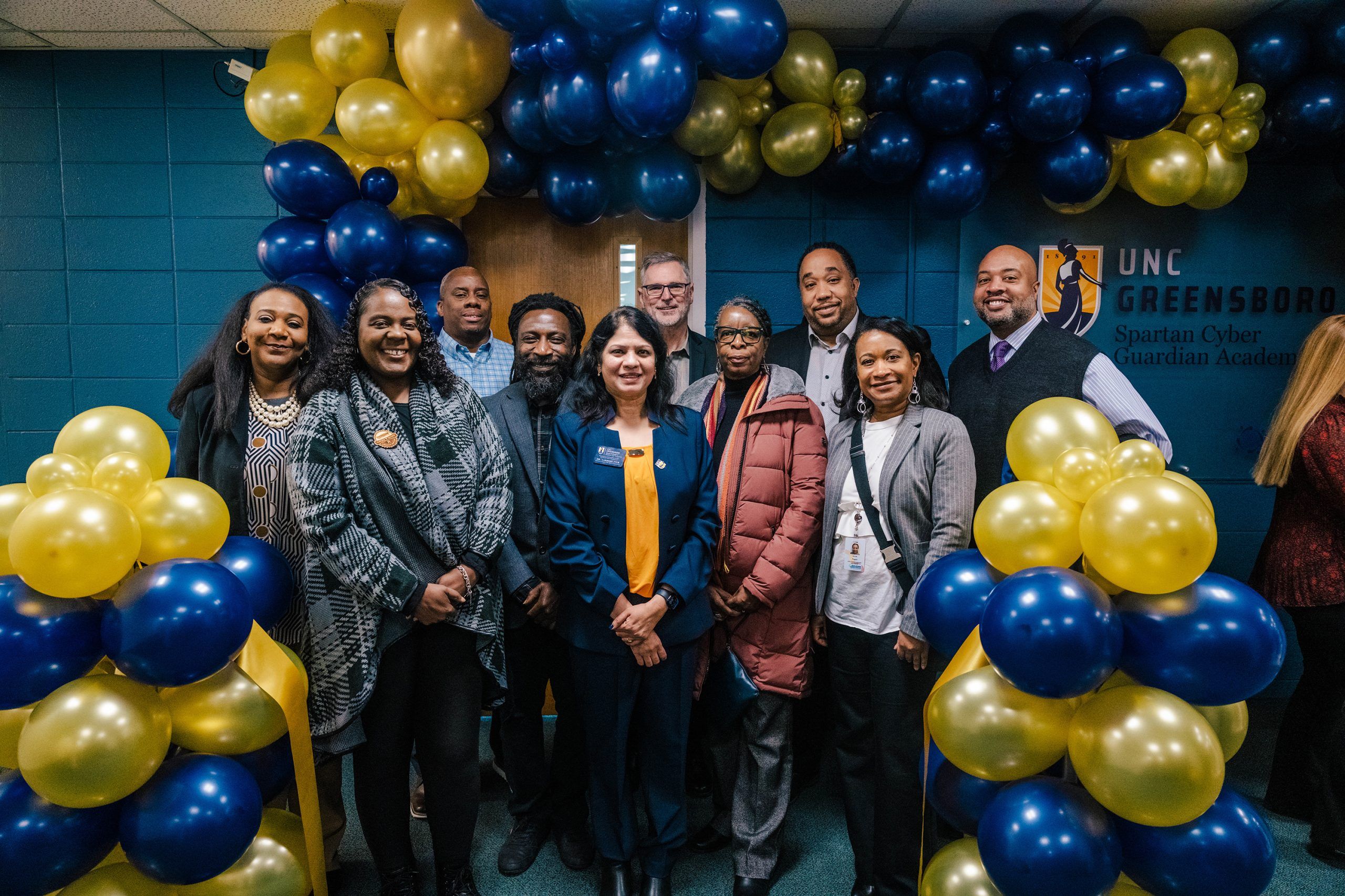 Community partners, students, faculty, and administrative leaders stand under an archway decorated with blue and gold balloons at an event. The wall behind them has the UNC Greensboro logo and text reading “Spartan Cyber Security Academy.”