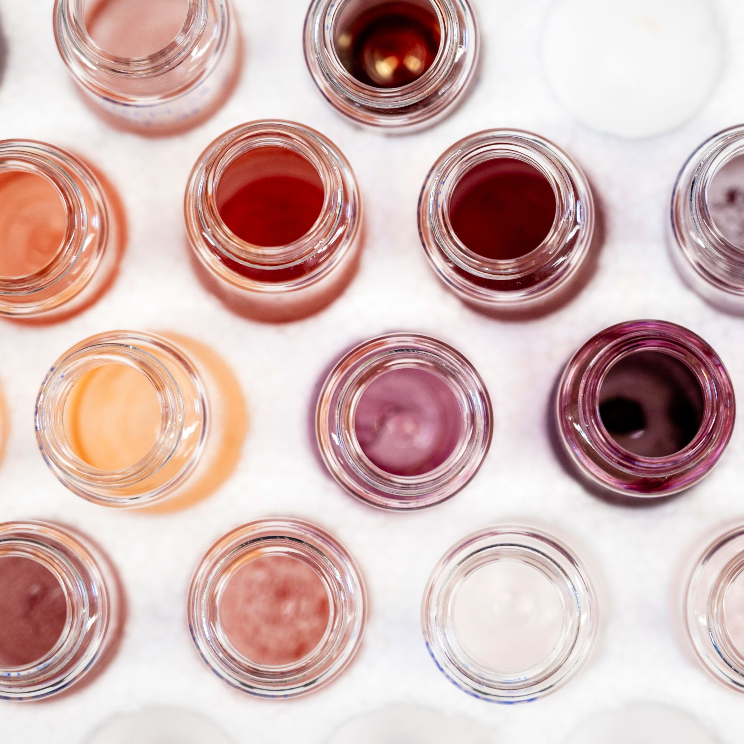 Top view of several small open glass jars arranged in a grid, each containing different shades of colored liquid or cream, ranging from peach and pink to red, purple, and clear, set against a white background.
