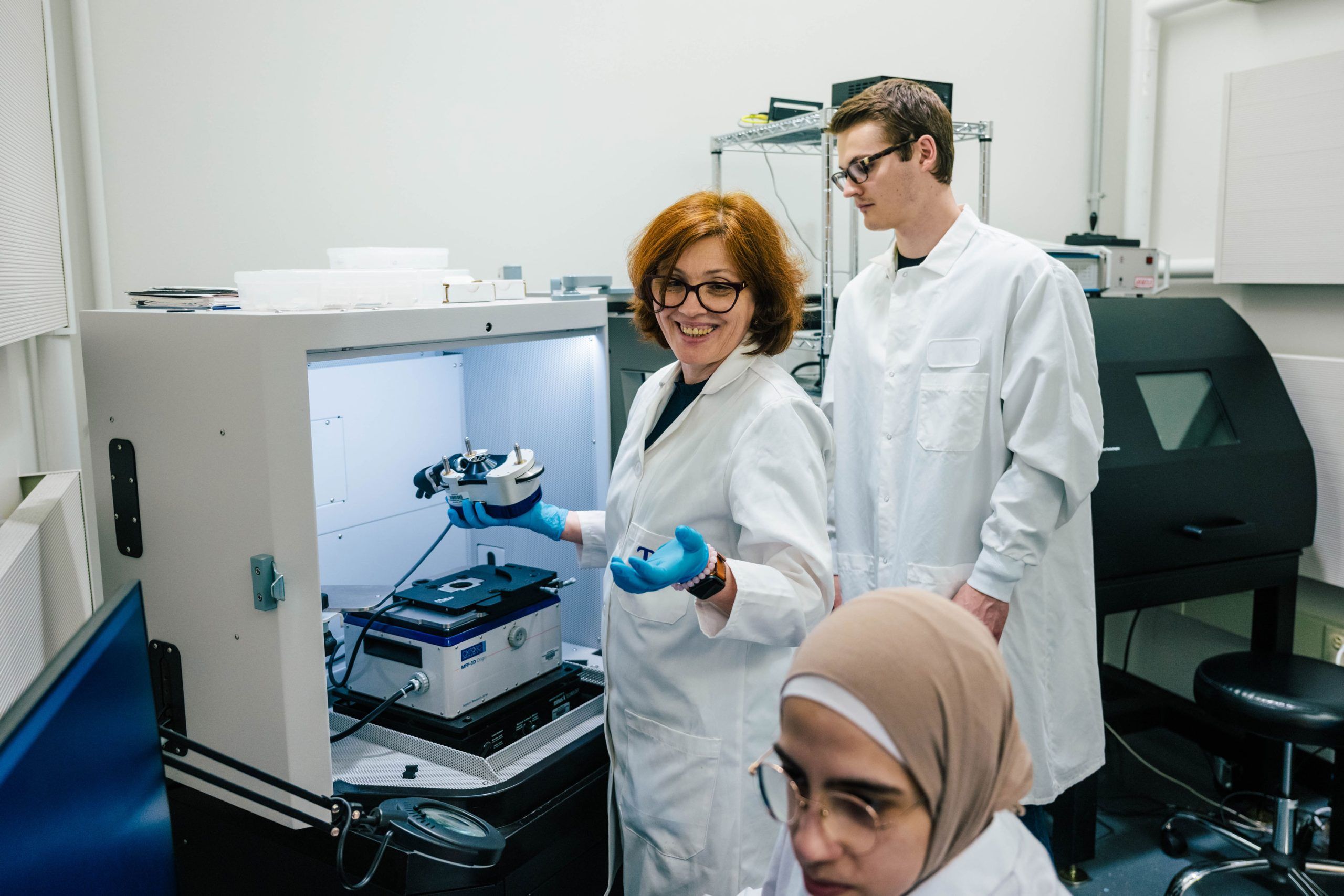 Dr. Ignatova and two of her students wearing white lab coats work in a laboratory. One person is holding a piece of equipment inside a large white enclosure with a microscope stage, while another stands nearby observing. A third person is seated in the foreground. The background includes shelves with lab supplies and a black instrument enclosure, creating a clean, organized research environment.
