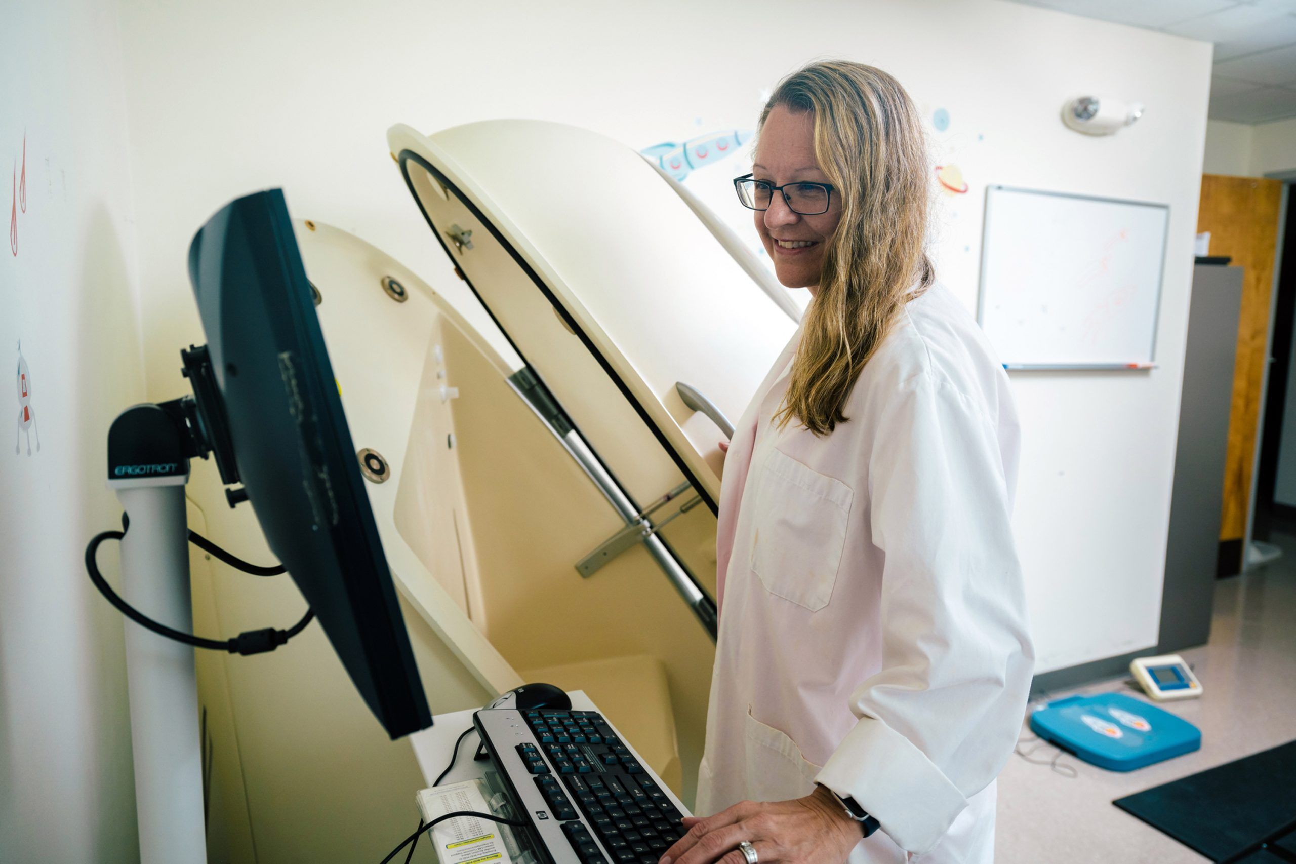 Laurie Wideman in a lab coat operating a computer next to an open Bod Pod chamber used for body composition testing. A whiteboard and scale are visible in the background.