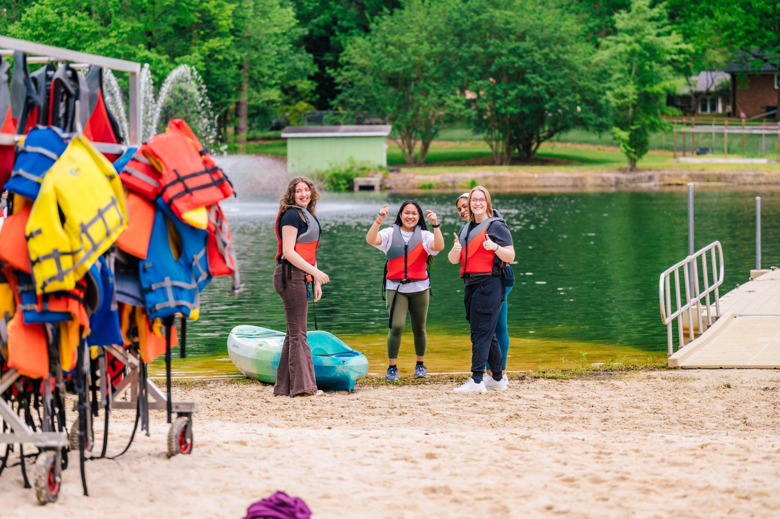 Three people wearing red life jackets stand on a sandy beach near a green kayak, preparing for a water activity. A rack of colorful life jackets is visible on the left, and the background shows a calm lake with a fountain, a dock, and green trees.
