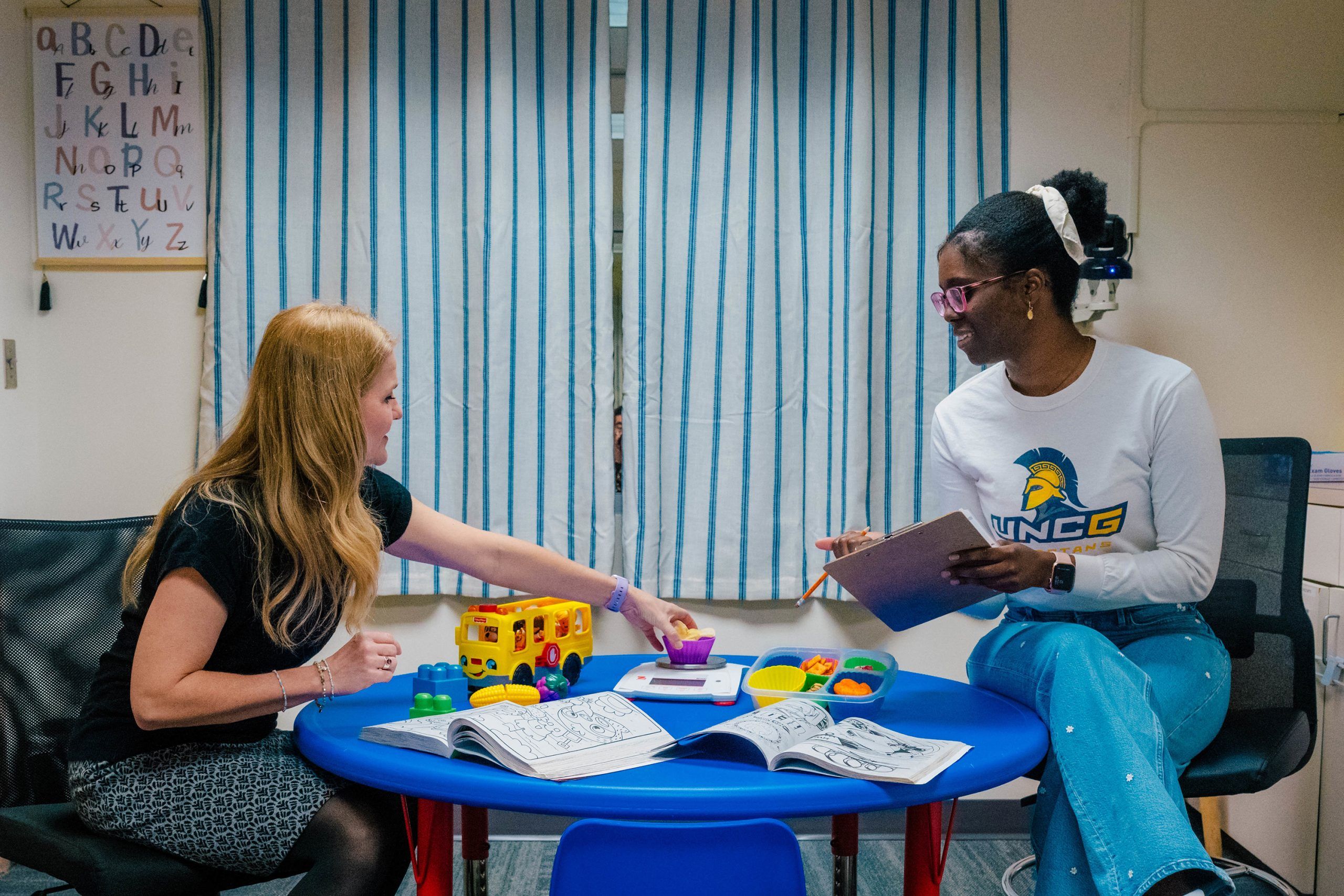 Dr. Lenka Shriver and undergrad Jahleen Gourdine seated at a small blue table in a classroom setting with striped curtains in the background; the table holds open workbooks, colorful educational toys, and a clipboard, while an alphabet chart hangs on the wall.