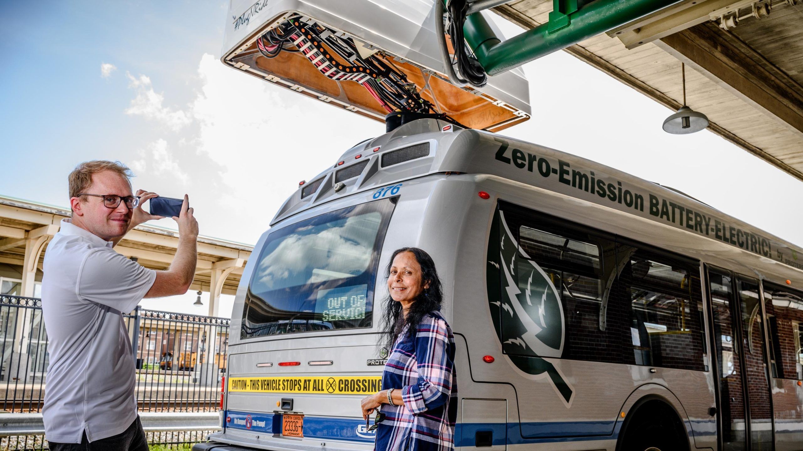 Dr. Sultana stands near a silver zero-emission battery electric bus at a charging station, while another person takes a photo using a smartphone. The bus has text reading “Zero-Emission BATTERY ELECTRIC” along the side and is positioned under a large overhead charging unit connected by cables. The setting appears to be an outdoor transit facility with a metal fence and station structures visible in the background.