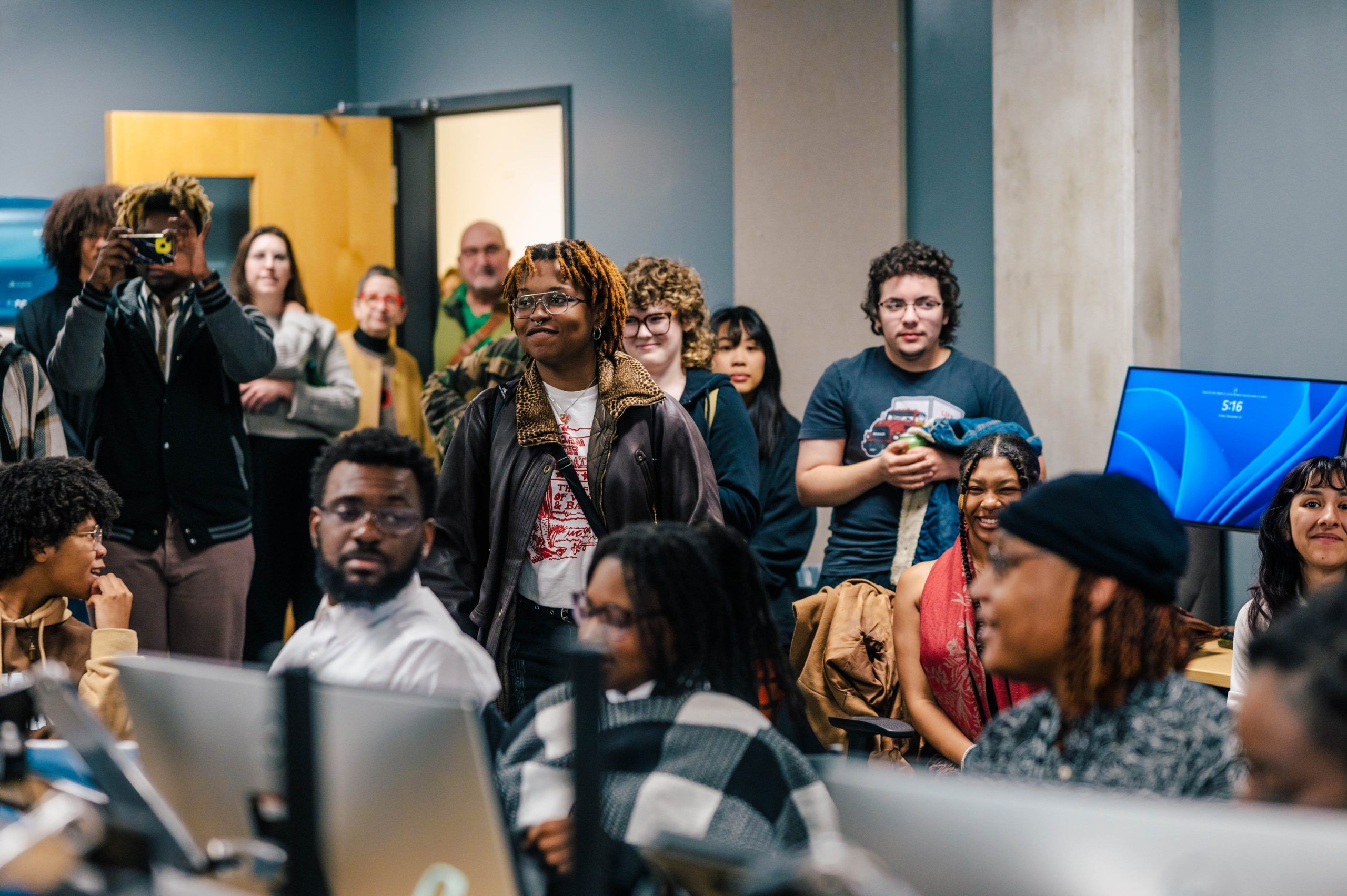 A crowded classroom or computer lab with multiple people standing and sitting around workstations. Several desktop monitors are visible, including one displaying a blue login screen. The setting appears to be an academic or training environment, with a mix of individuals gathered for a presentation or group activity.