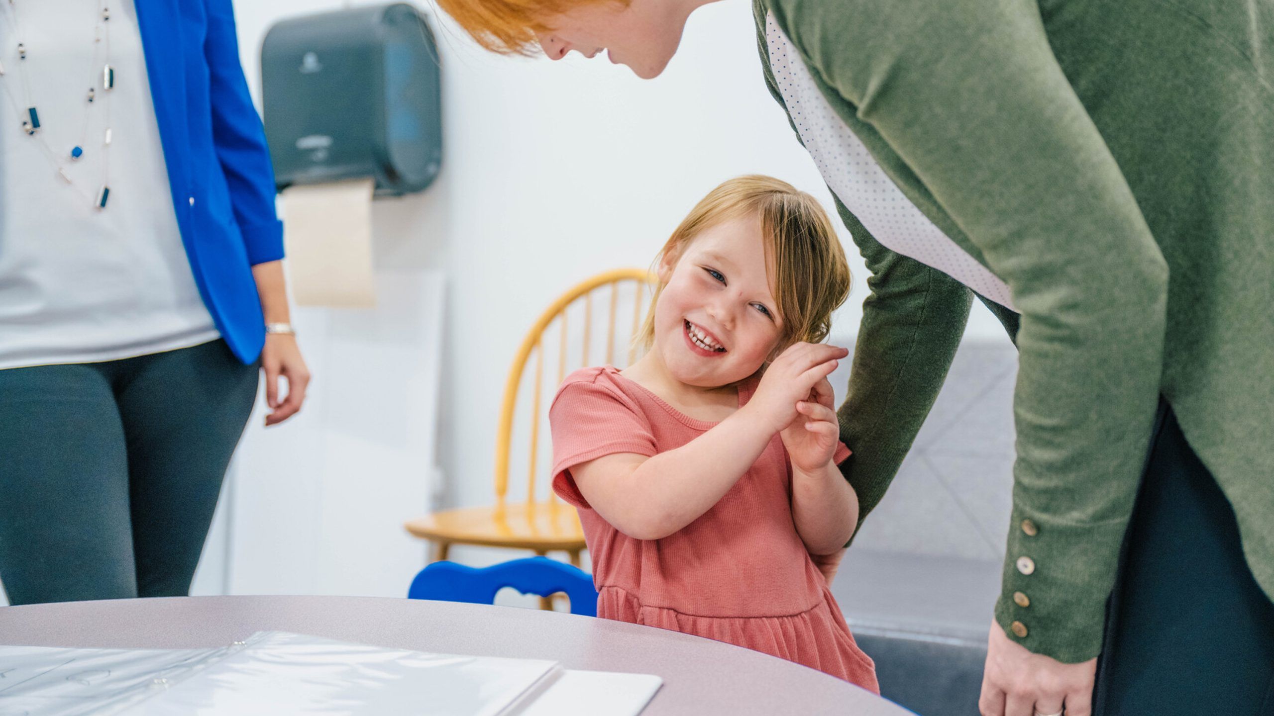Child in a pink dress standing near a table with a binder, while two adults interact with the child in a brightly lit room. A wooden chair and wall-mounted paper towel dispenser are visible in the background.
