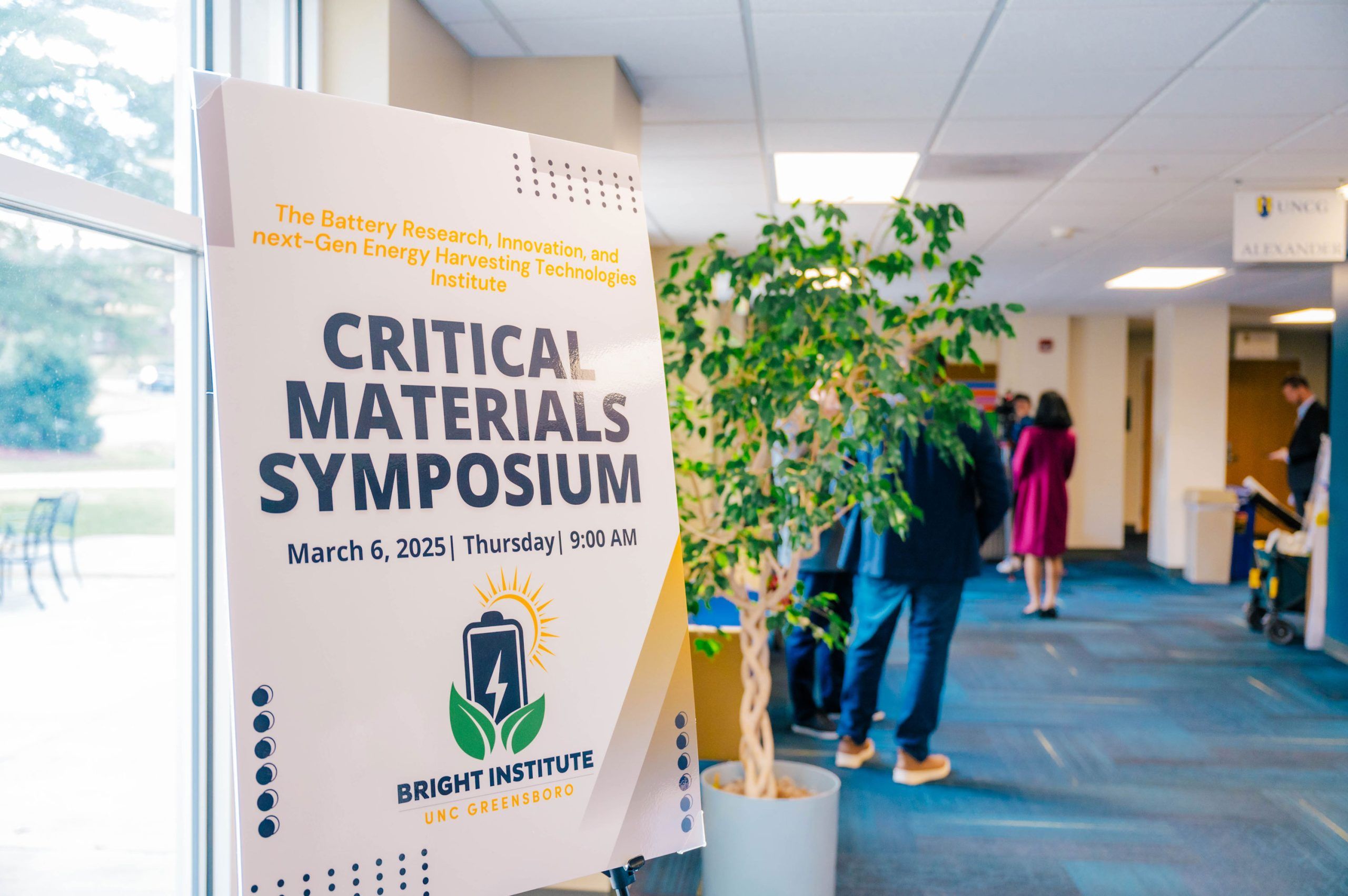 A large poster on an easel in a hallway announces the “Critical Materials Symposium” hosted by the Bright Institute at UNC Greensboro. The text includes the date and time: March 6, 2025, Thursday, 9:00 AM. The poster features a logo with a battery icon and green leaves. In the background, people are gathered near a doorway, and a potted plant is visible beside the sign.