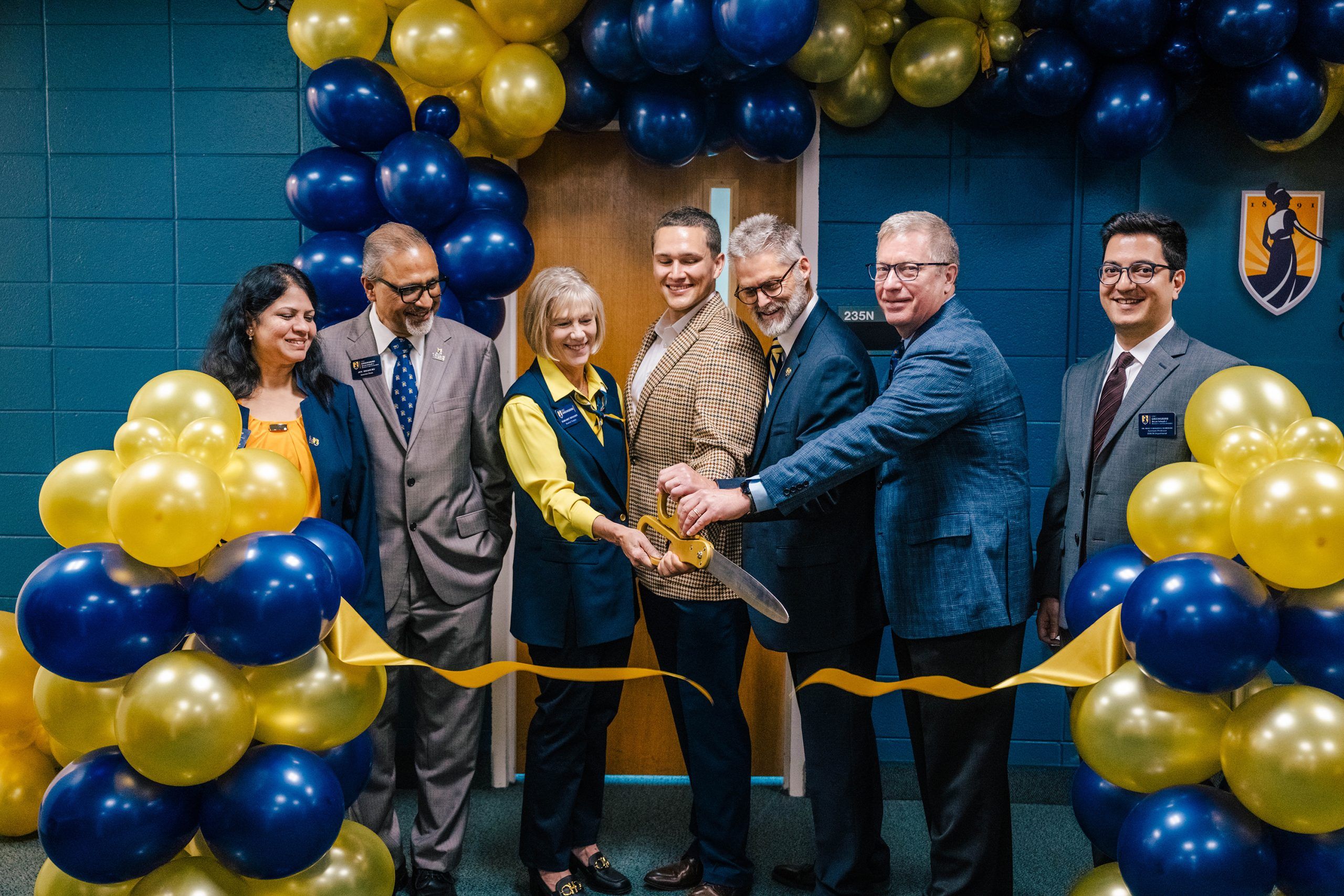 A ribbon-cutting ceremony with several people standing under an arch of blue and gold balloons. Two individuals hold large scissors cutting a gold ribbon stretched across the doorway. The UNC Greensboro logo is visible on the wall in the background.