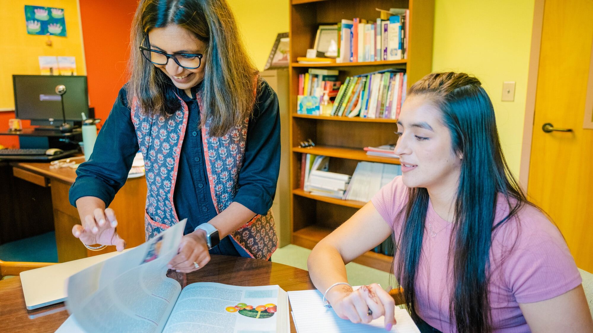 Jigna Dharod and Selena Villa seated at a desk in an office with bookshelves in the background; one is flipping through a textbook while the other writes in a notebook.