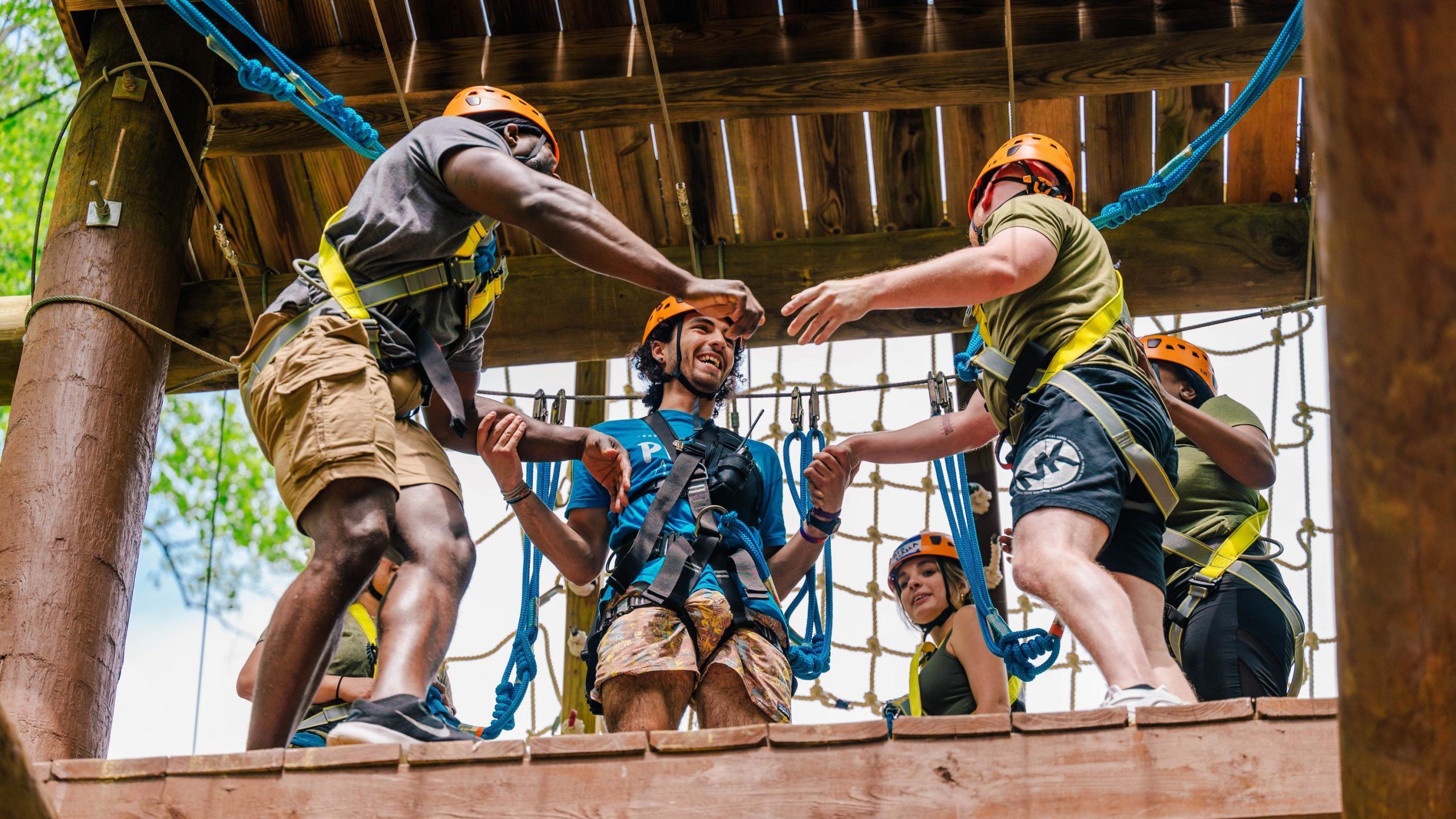 Several people wearing safety harnesses and orange helmets participate in a ropes course activity on a wooden platform. They are helping each other navigate through a section with rope netting, using handholds and support ropes. The setting is outdoors, with green foliage visible in the background, and the structure is made of wood with blue climbing ropes attached.
