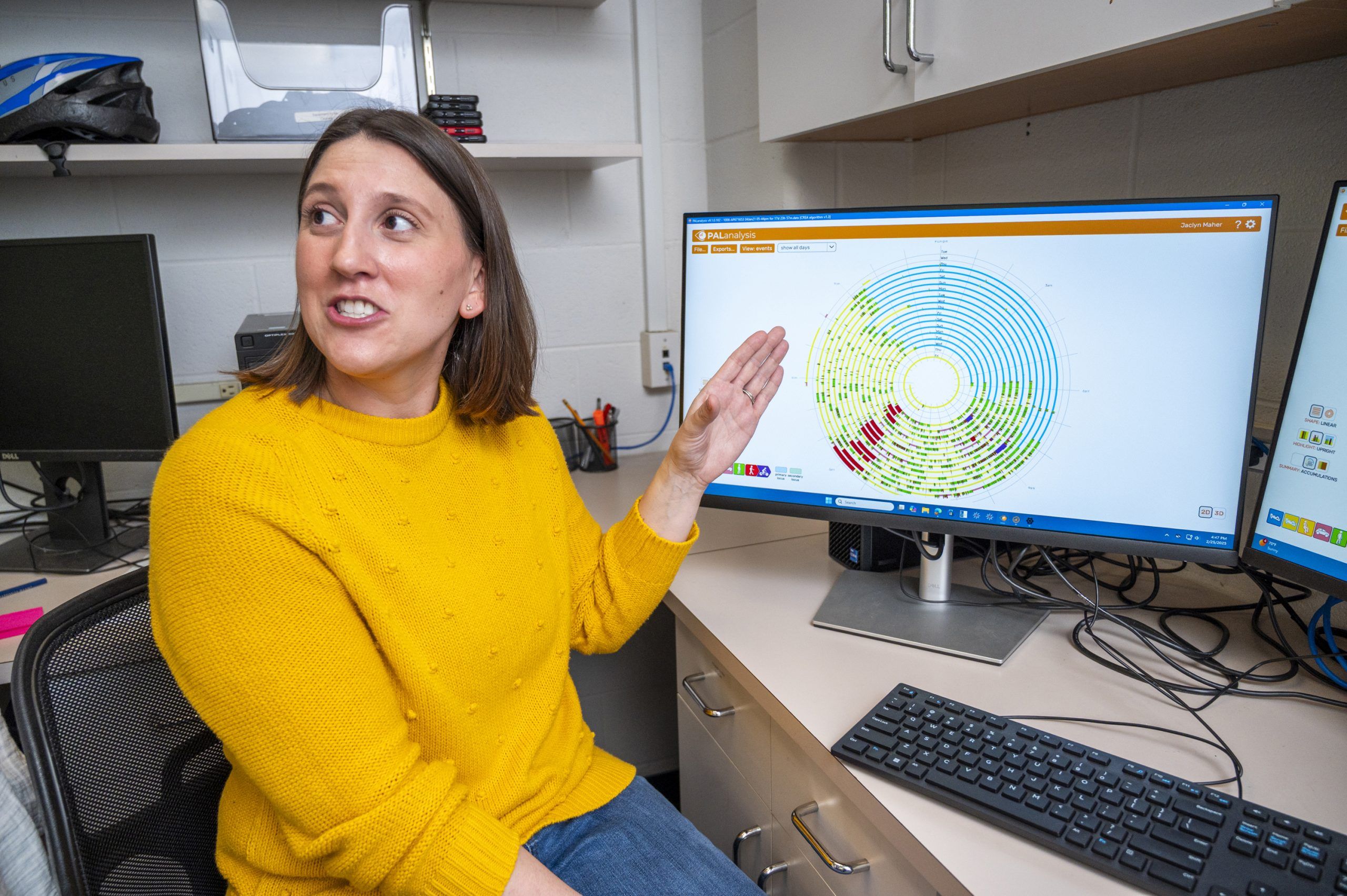 Dr. Maher wearing a bright yellow sweater sits at a desk in an office and gestures toward a computer monitor displaying a circular chart with multiple concentric rings in various colors, including green, yellow, and red. The desk has a keyboard, mouse, and cables, and another monitor is partially visible to the side. Shelves and office equipment are in the background.