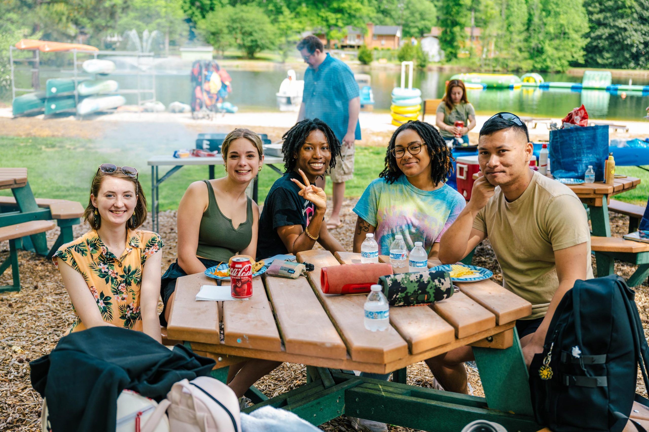 A group of people sit at a wooden picnic table outdoors near a lake, with kayaks and paddle boats visible in the background. The table has water bottles, snacks, and a soda can on it, and a chessboard is placed in the center. Trees and recreational equipment surround the area, creating a casual, scenic setting.