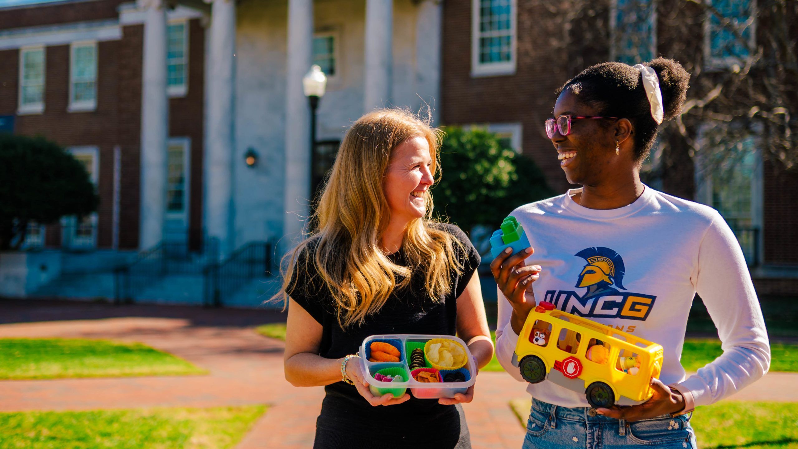 Dr. Shriver and Jahleen Gourdine stand outside in front of a brick building with white columns, holding educational items. Dr. Shriver holds a divided container with colorful toy fruits and vegetables, while Gourdine holds a yellow toy school bus and a green block. The setting is a sunny day on a campus lawn with a paved walkway and trees in the background.