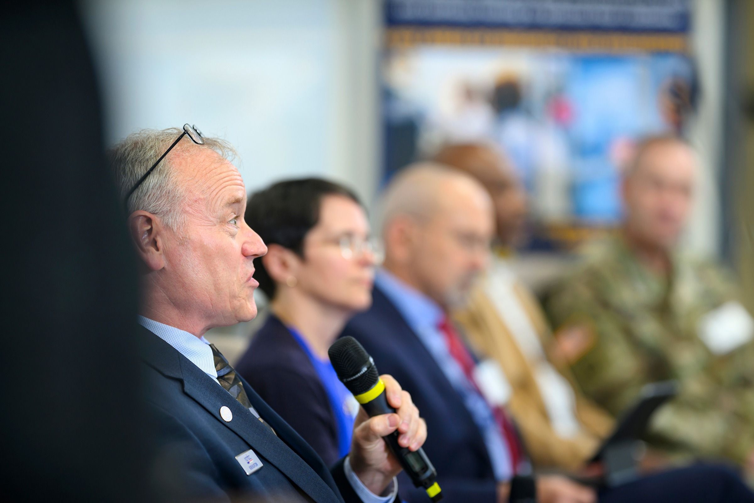 A person in a suit holds a microphone during a panel discussion or event. Several other attendees are seated in the background, including one in a military uniform. The setting appears to be a formal symposium or conference, with a banner featuring text and images visible behind the group.