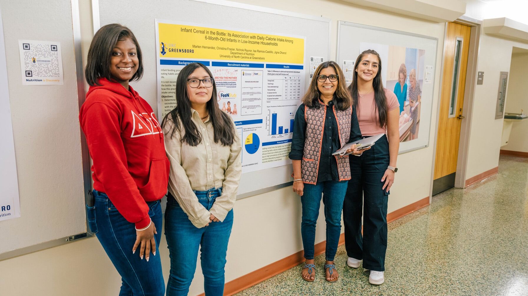 Jigna Dharod, Selena Villa, and other undergraduate nutrition students standing in a hallway next to a large research poster mounted on a bulletin board; the poster includes charts, graphs, and text about infant cereal and feeding practices.