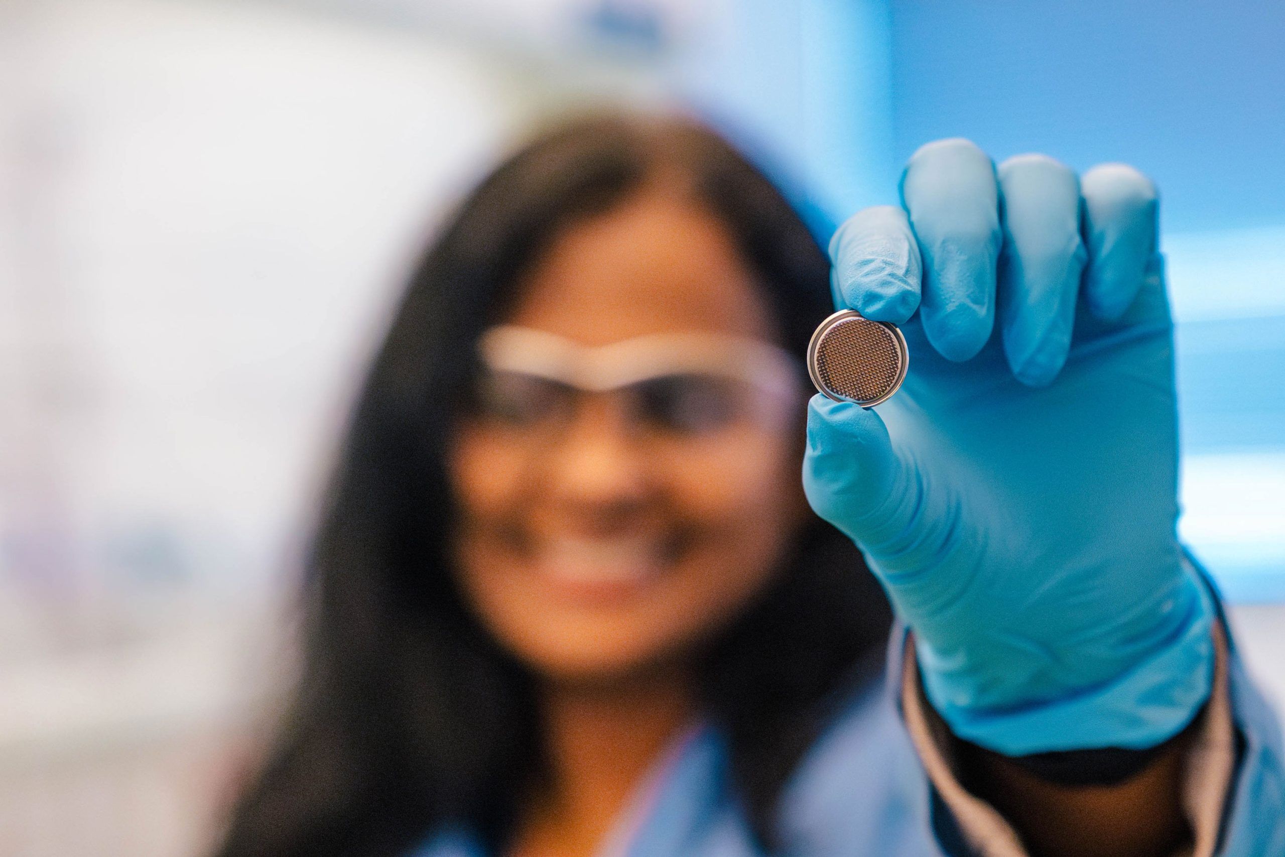 Dr. Rathnayake's gloved hand holds a small, round metallic object resembling a coin cell battery or sensor component. The object has a textured or mesh-like surface on one side. The background is softly blurred, showing a laboratory setting with cool blue tones.