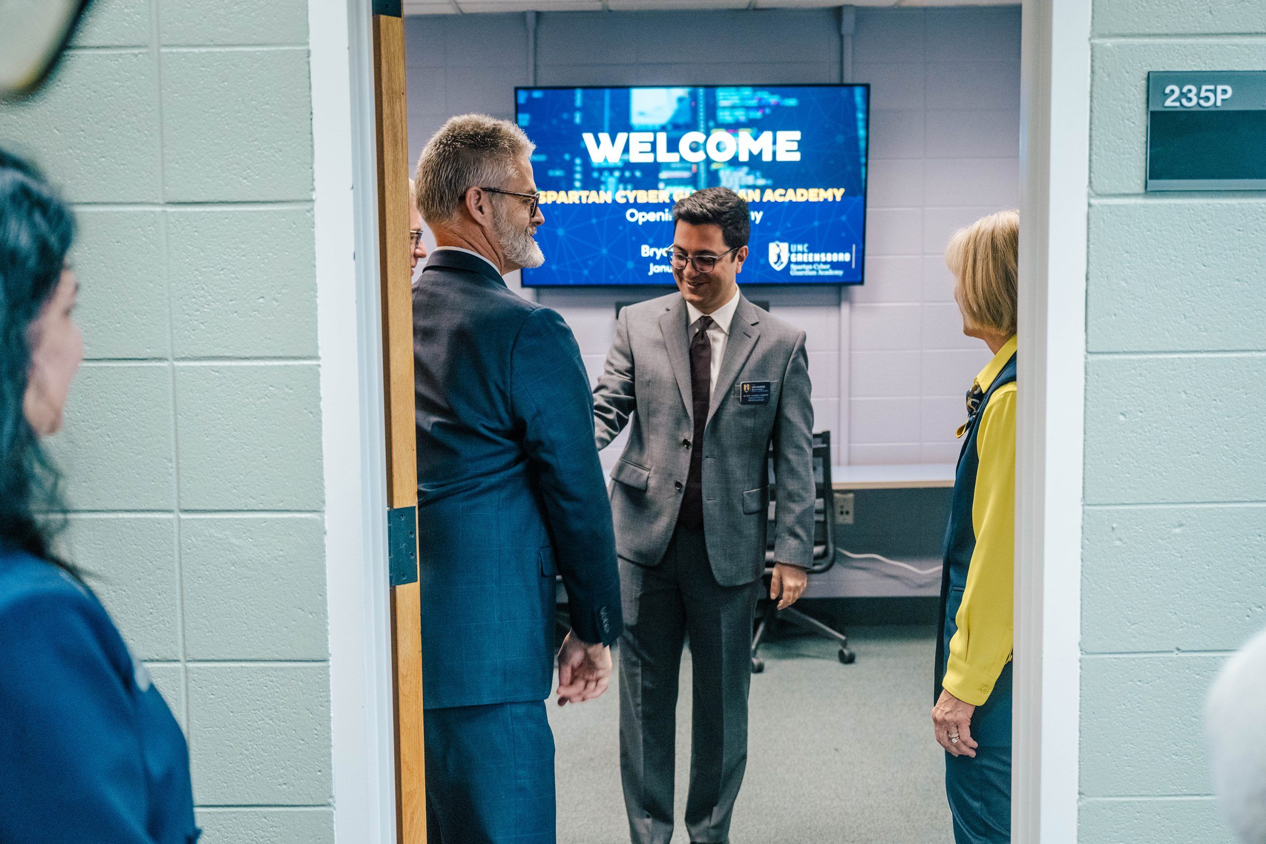 Three people stand in a doorway near a wall-mounted monitor displaying the message “WELCOME Spartan Cyber Security Academy Opening Ceremony” with the UNC Greensboro logo. The setting appears to be an office or classroom with gray walls and a sign reading “235P” on the door frame.