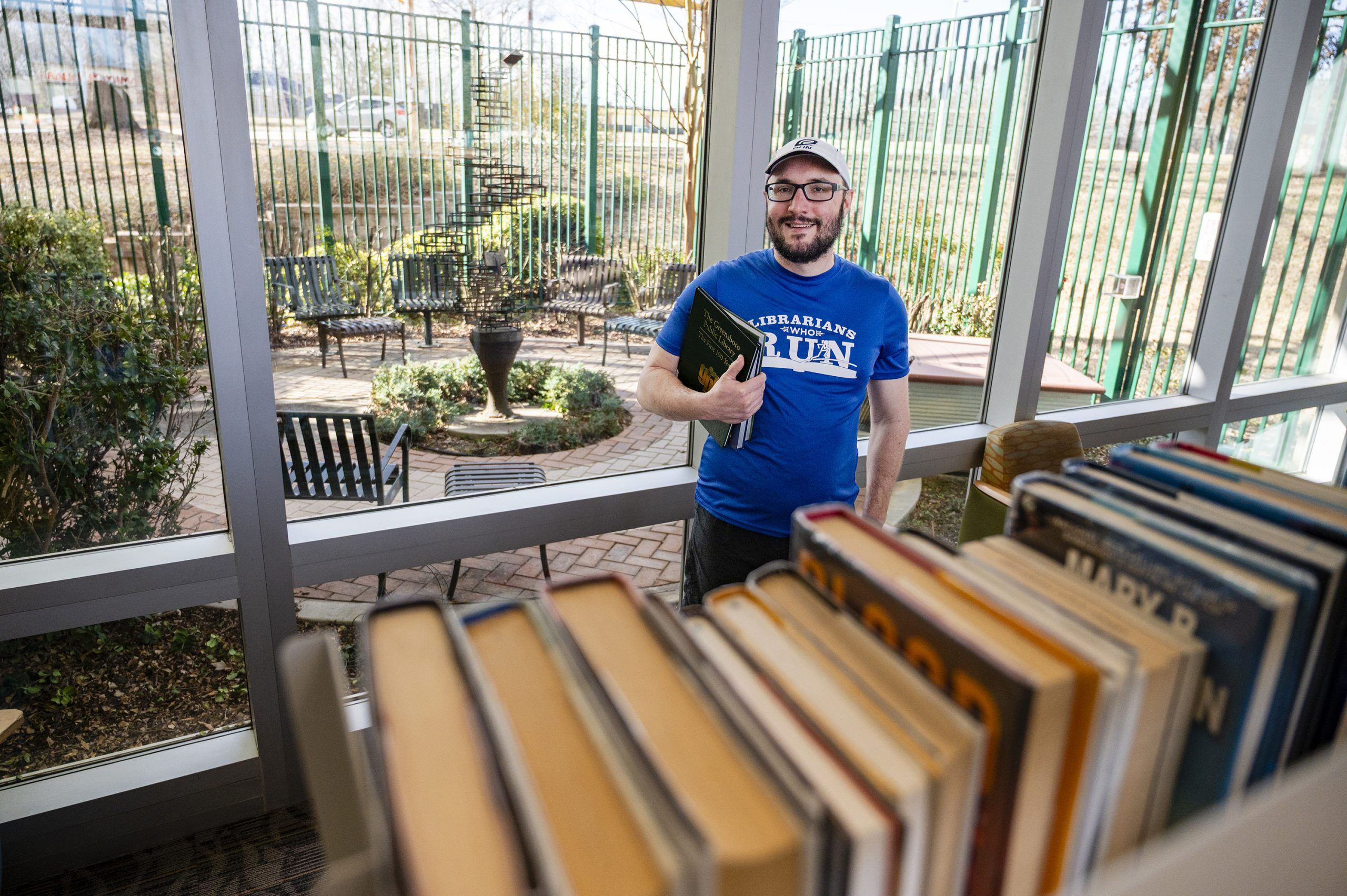 Dr. Noah Lenstra holds a book as he stands in front of a window and behind a shelf of books