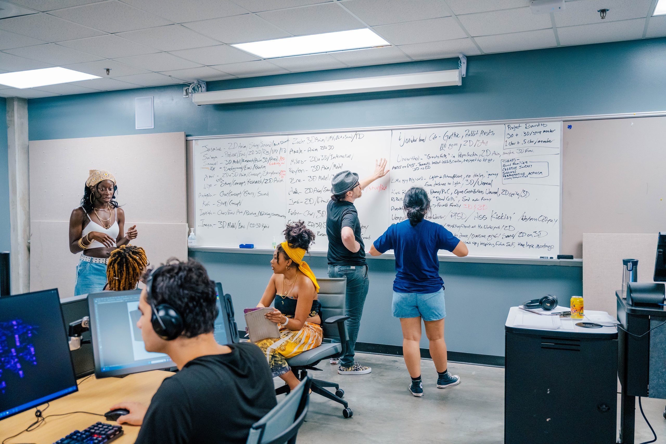 A classroom setting where several people are working collaboratively. Two individuals are writing on a large whiteboard filled with notes and lists, while others sit at desks with computers and notebooks. One person is standing and holding a tablet, and another is seated wearing headphones at a computer station. The room has gray walls, a tiled ceiling, and fluorescent lighting, creating an academic or workshop environment.
