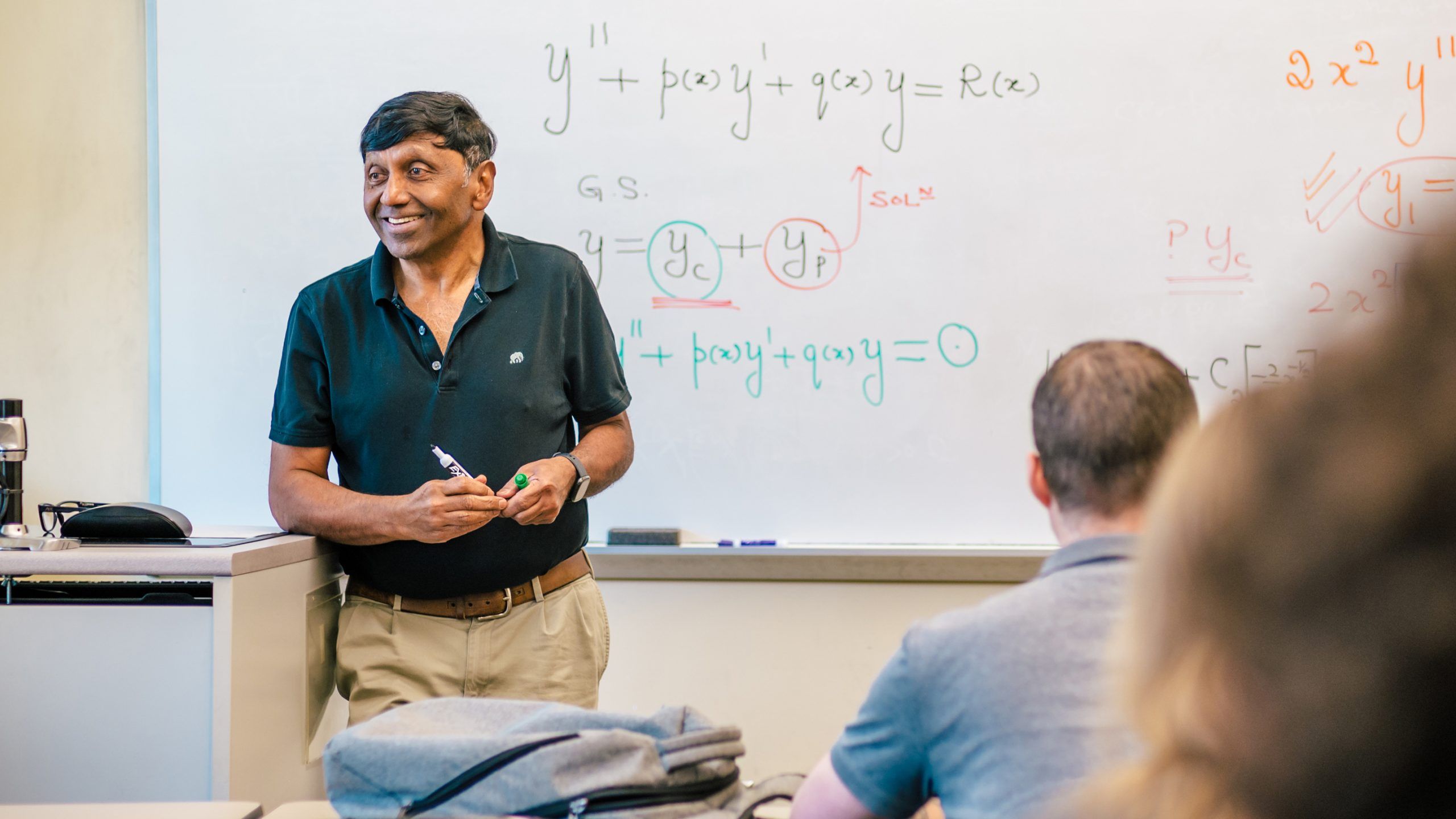 Dr. Shivaji standing at the front of a classroom near a whiteboard with mathematical equations, addressing students seated at desks.
