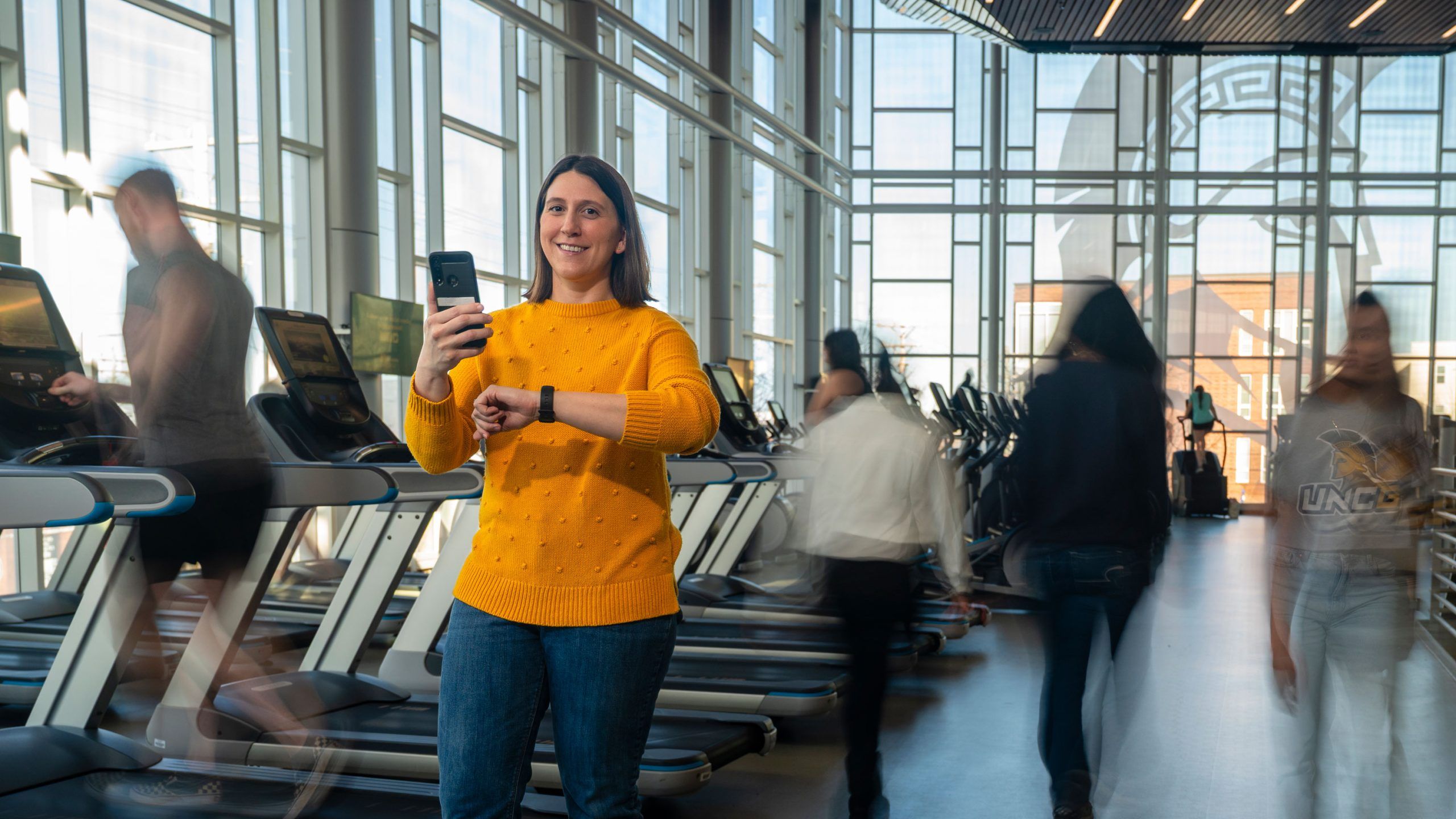 Dr. Maher in a bright yellow sweater standing in a fitness center, checking a smartwatch while holding a phone, with treadmills and people exercising in the background.