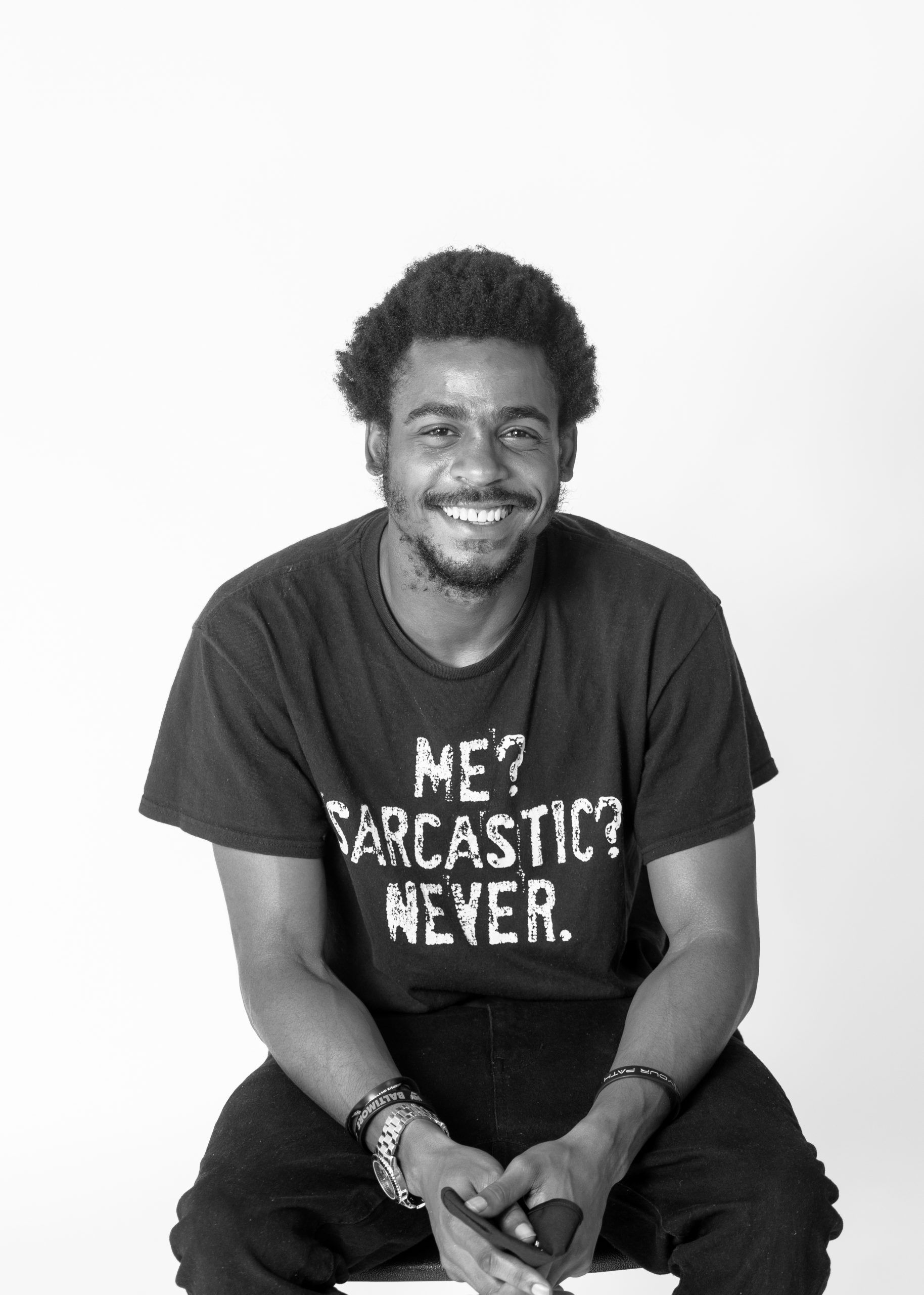A black and white photo of a Black man smiling wearing a t-shirt that says "Me? Sarcastic? Never."