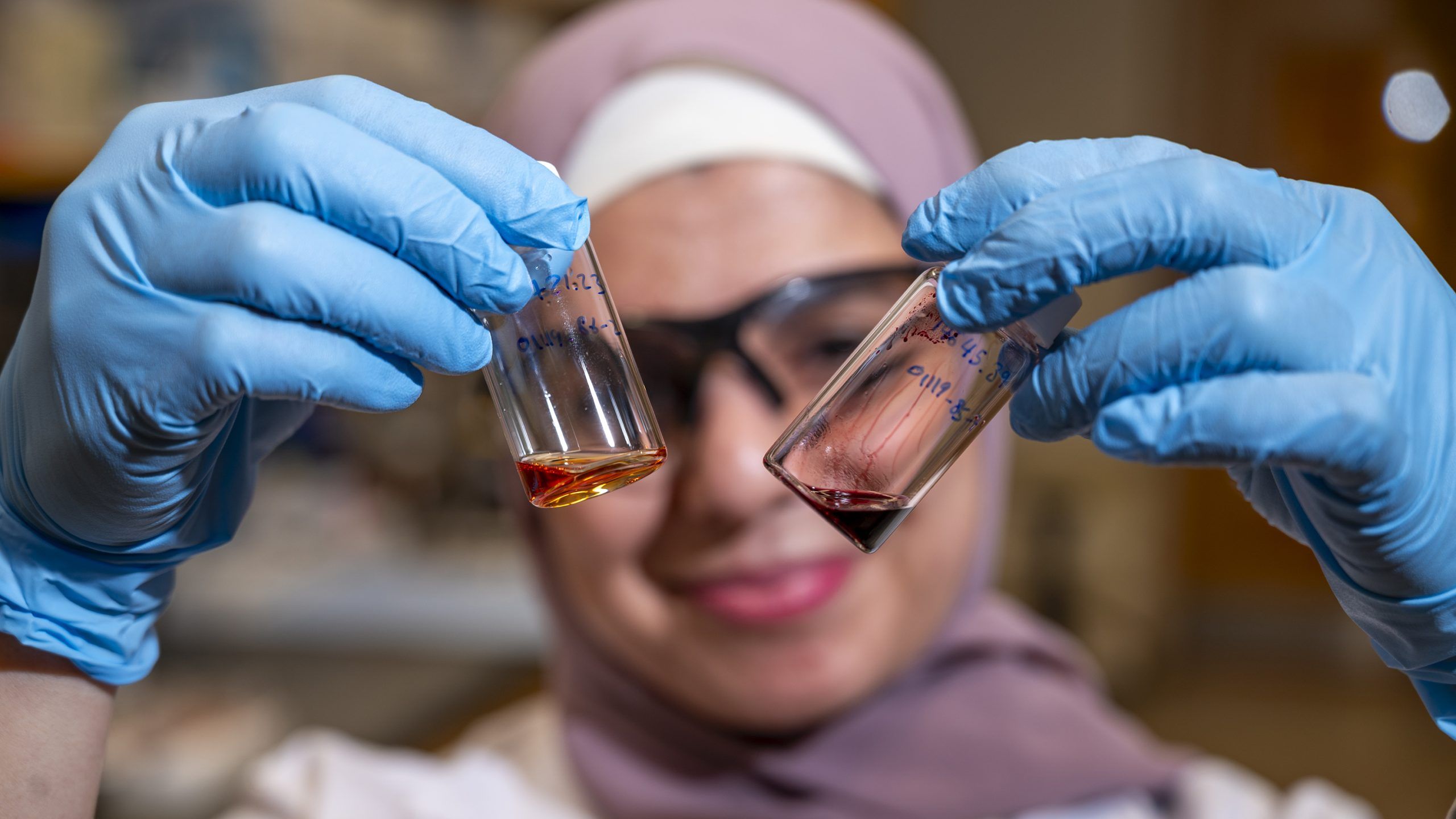 Ph.D. student Reema Al-Qiam wearing blue gloves holding a small laboratory vial or sample tube at eye level for examination.