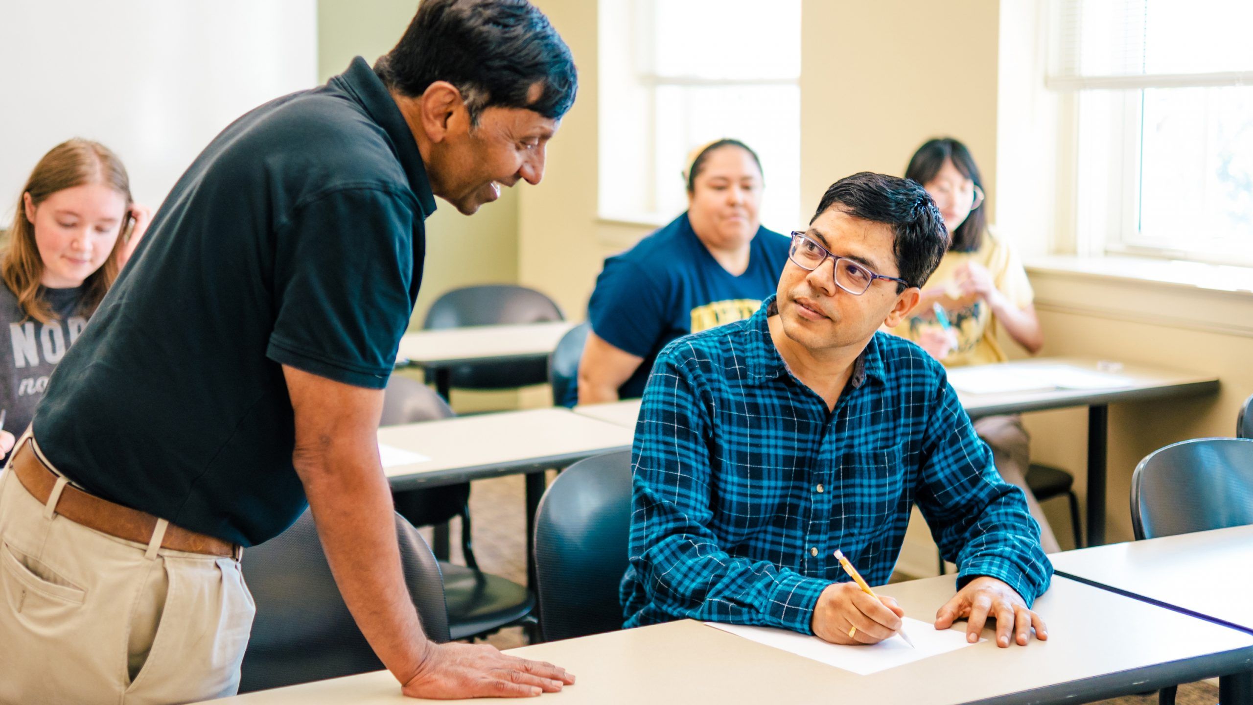 Dr. Shivaji leans over a classroom desk, speaking with a student who looks up and listens while holding a pencil and paper.