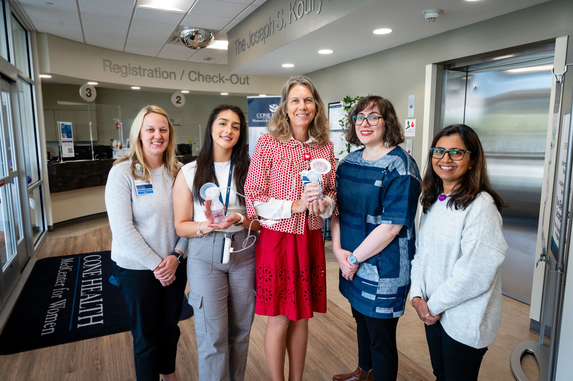 Cone Health's Marjorie Jenkins, grad student Selene Villa, Julie Wenzel, Jasmine DeJesus, and Jigna Dharod standing in a hospital lobby near a registration desk. Background shows signage for registration and Cone Health MedCenter for Women.