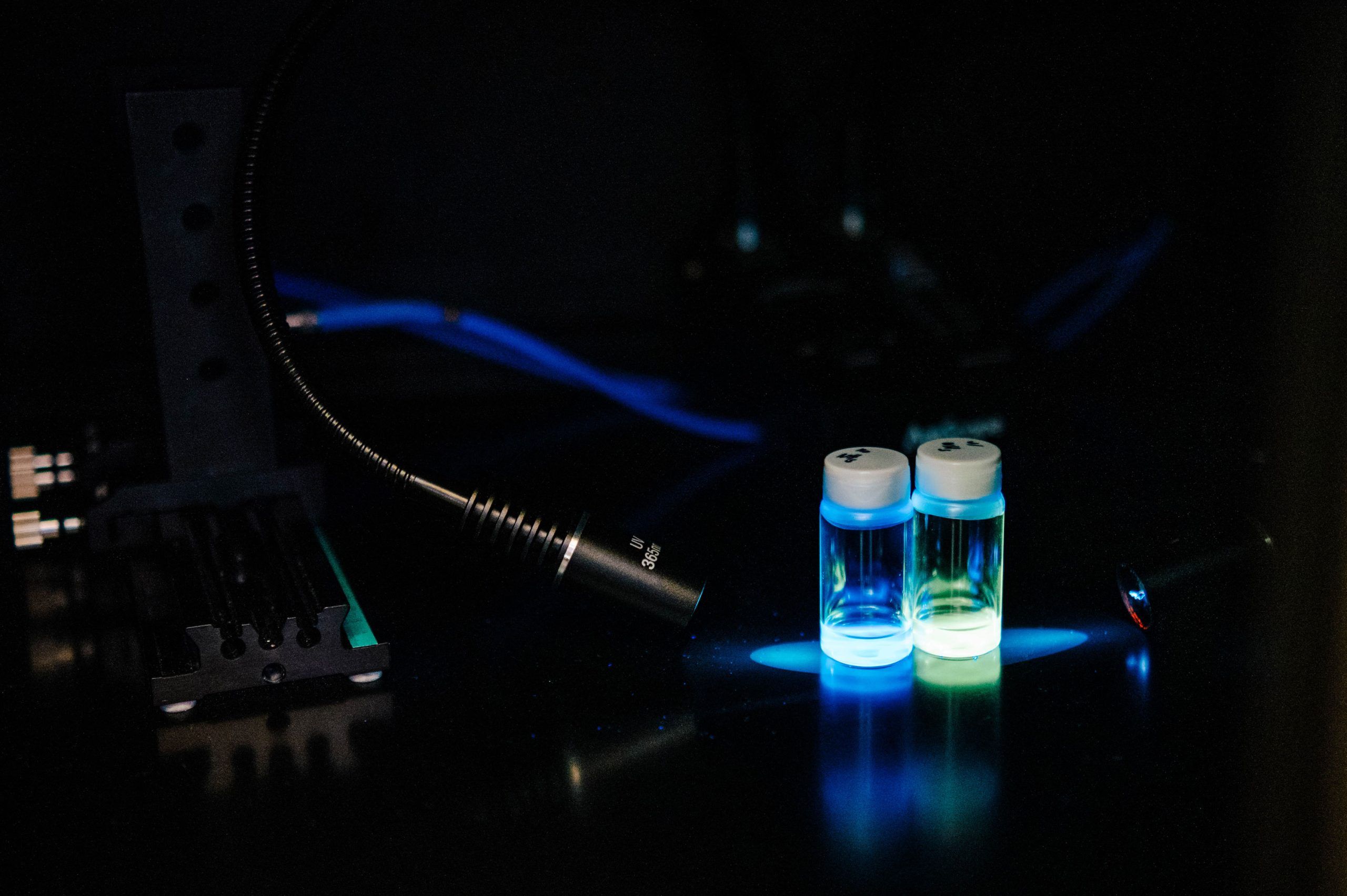 Two small glass vials with white caps containing glowing liquid—one blue and one green—illuminated under a focused light source on a dark laboratory surface. Nearby are a flexible lamp, cables, and a metal mounting bracket.