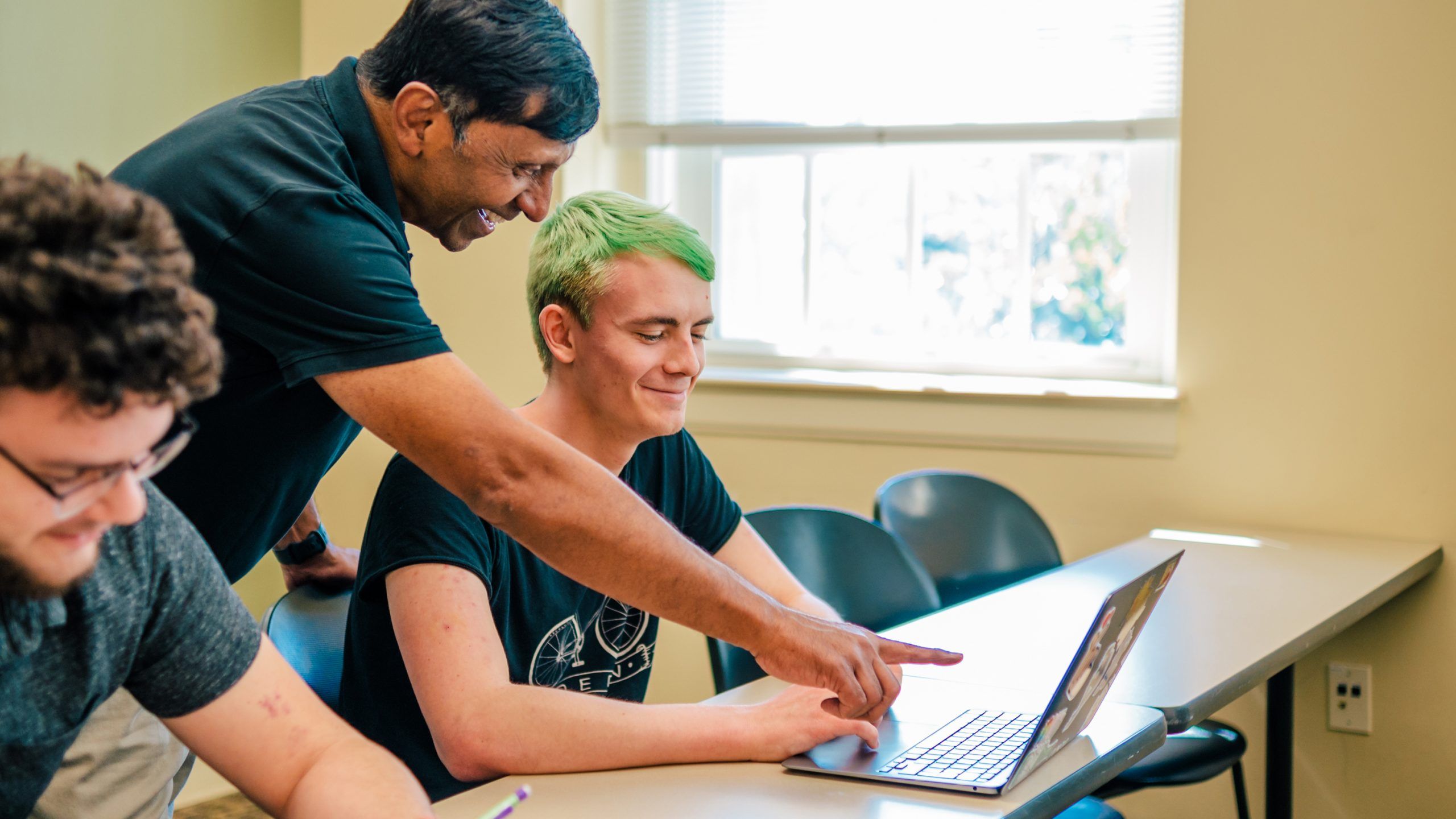 Dr. Shivaji leans over a student’s desk, smiling as he points to the student’s laptop screen during class.