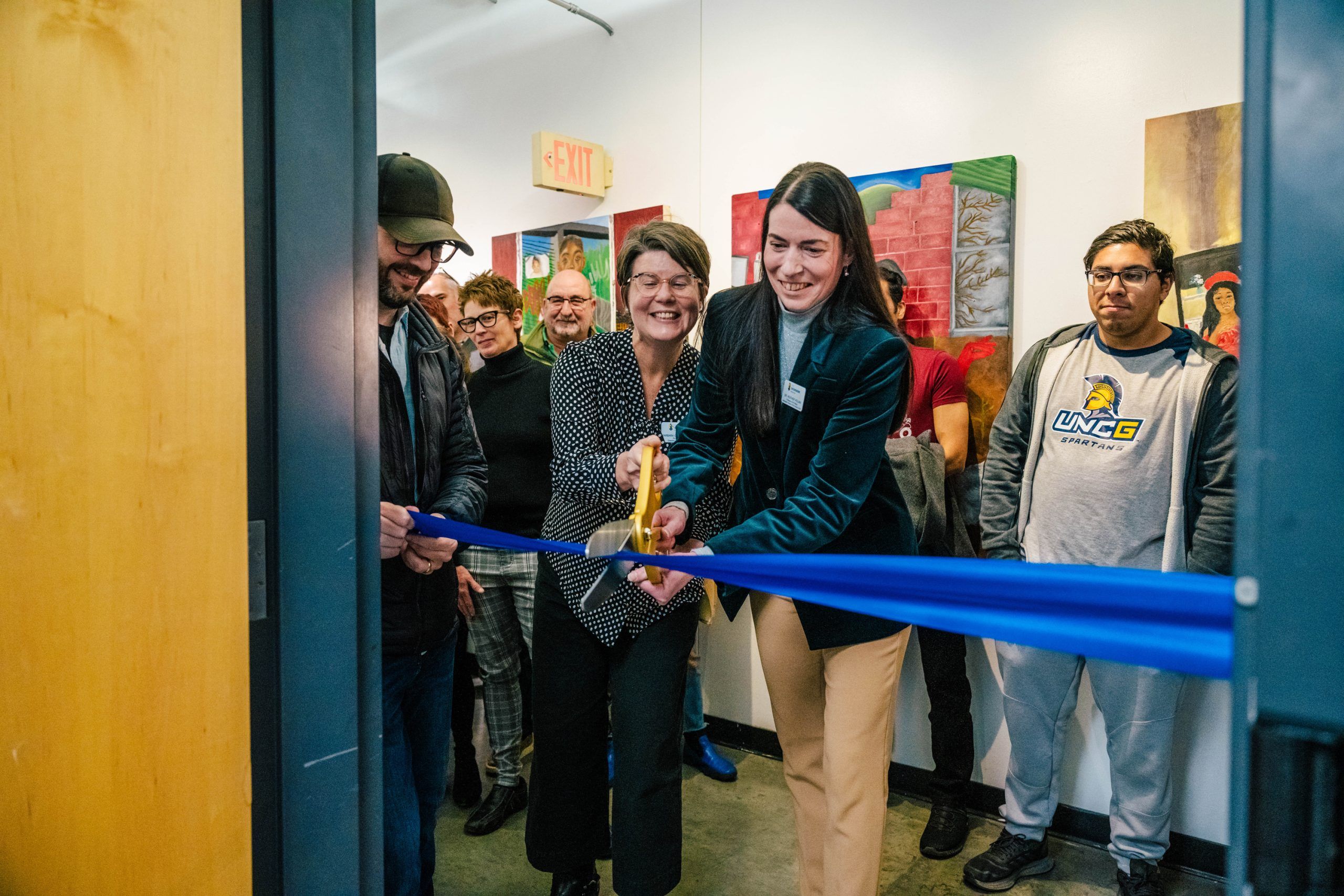 A ribbon-cutting ceremony inside an art gallery or exhibition space. Two individuals hold large scissors and cut a bright blue ribbon stretched across the doorway. Several people stand behind them, and colorful artwork is displayed on the walls in the background. An exit sign is visible above the door.
