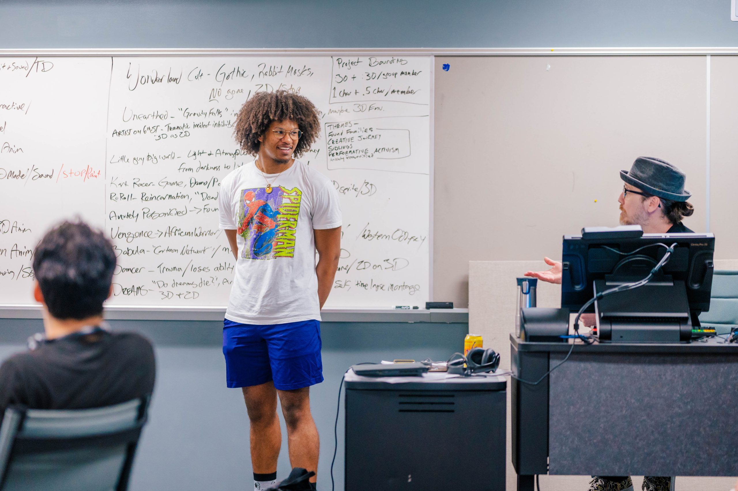 A classroom setting with a large whiteboard covered in handwritten notes and lists. A person stands in front of the board wearing a graphic T-shirt and blue shorts, while another person sits at a desk in the foreground and a third person is seated behind a computer station on the right. The room includes a gray wall, a desk with a computer monitor, and a small cabinet with items on top, creating an academic or workshop environment.