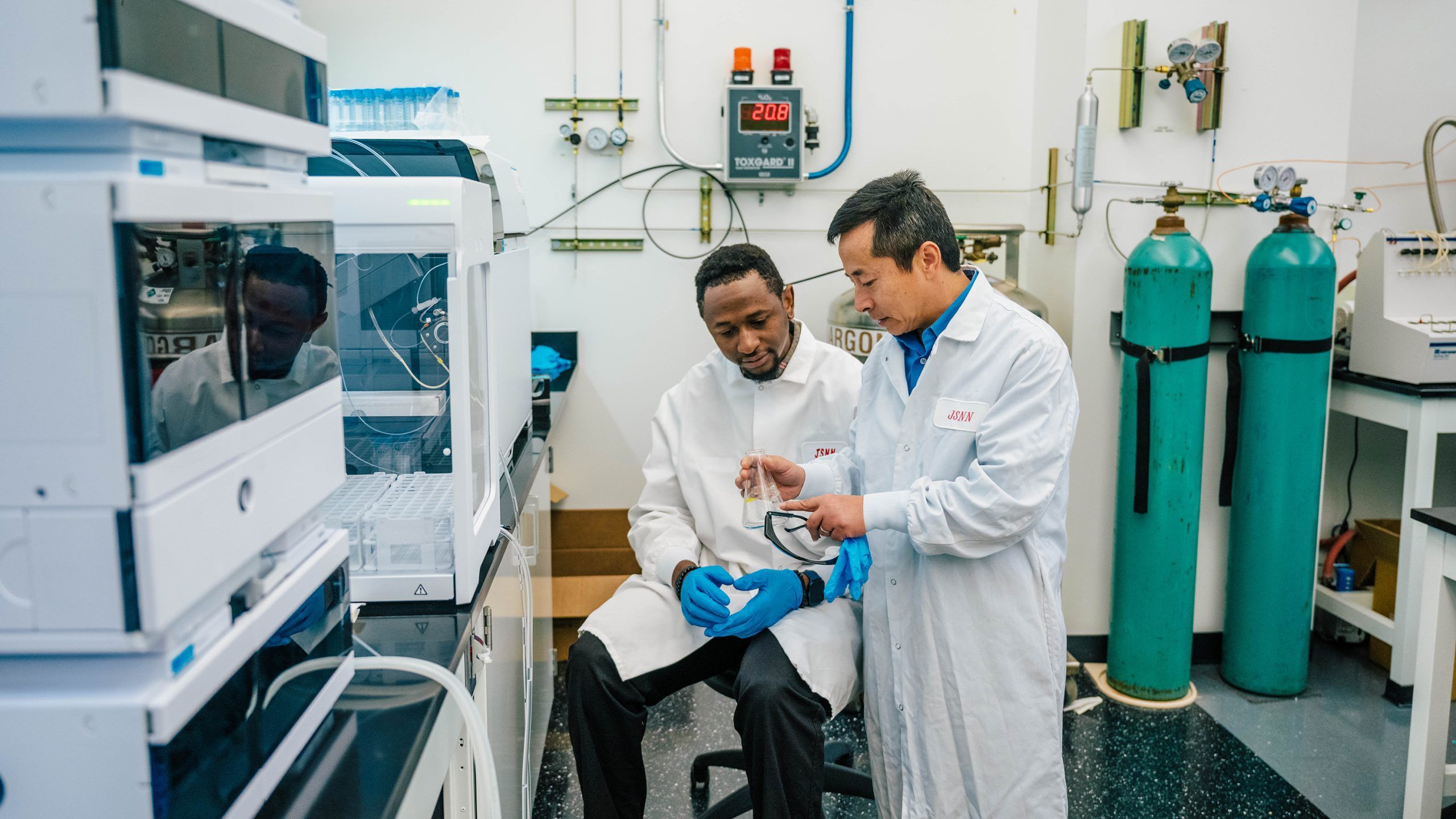 Dr. Wei and one of his students wearing lab coats and gloves work together in a laboratory. The student is seated while Dr. Wei holds a glass flask and explains something. Laboratory equipment, gas tanks, and instruments are visible around them.
