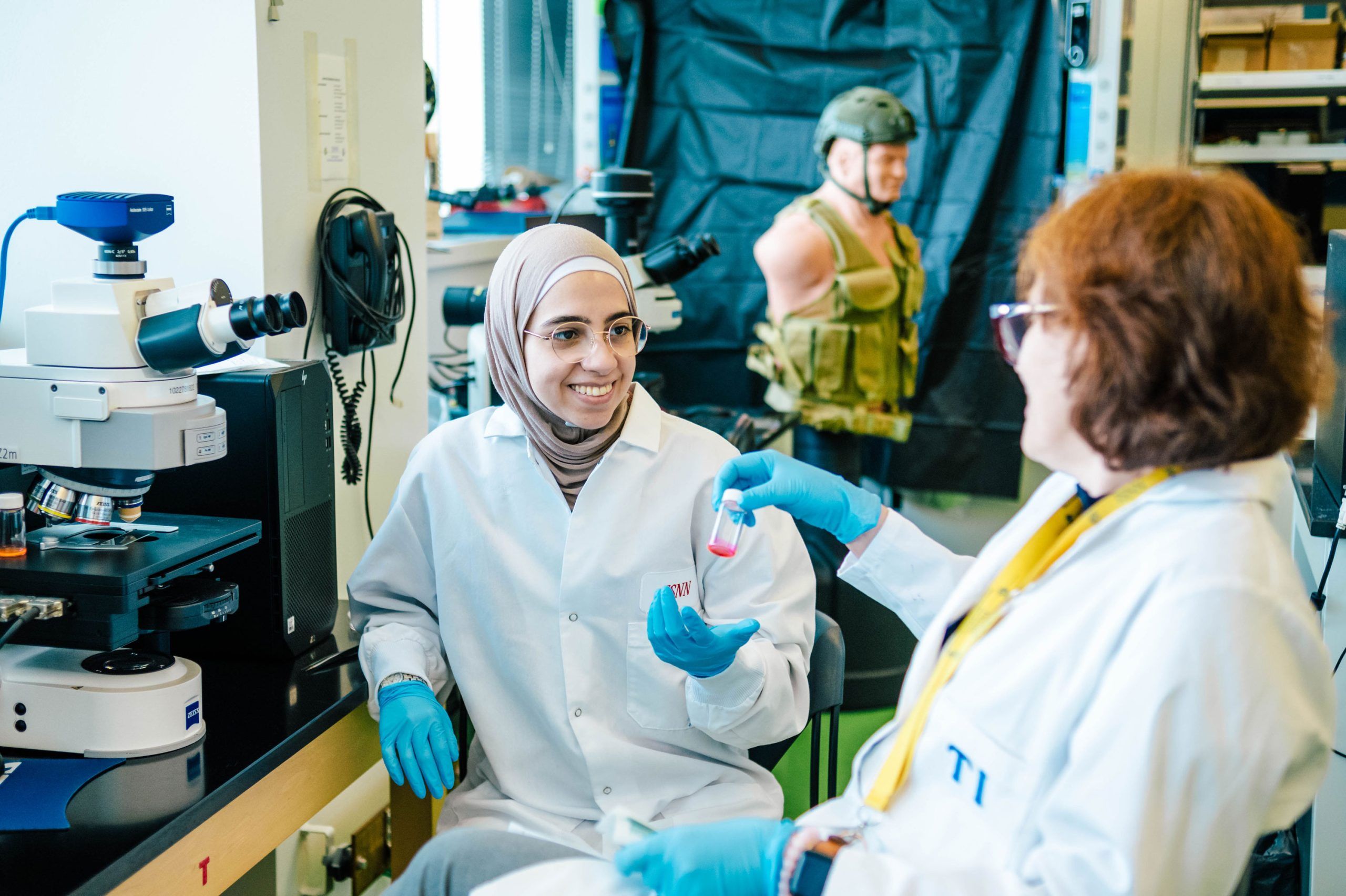 Dr. Ignatova and one of her students in a laboratory wearing white coats and blue gloves, seated near a microscope. Dr. Ignatova is holding a small vial with pink liquid while the student is gestures toward it. The background includes shelves with lab supplies and a mannequin torso wearing a green tactical vest.