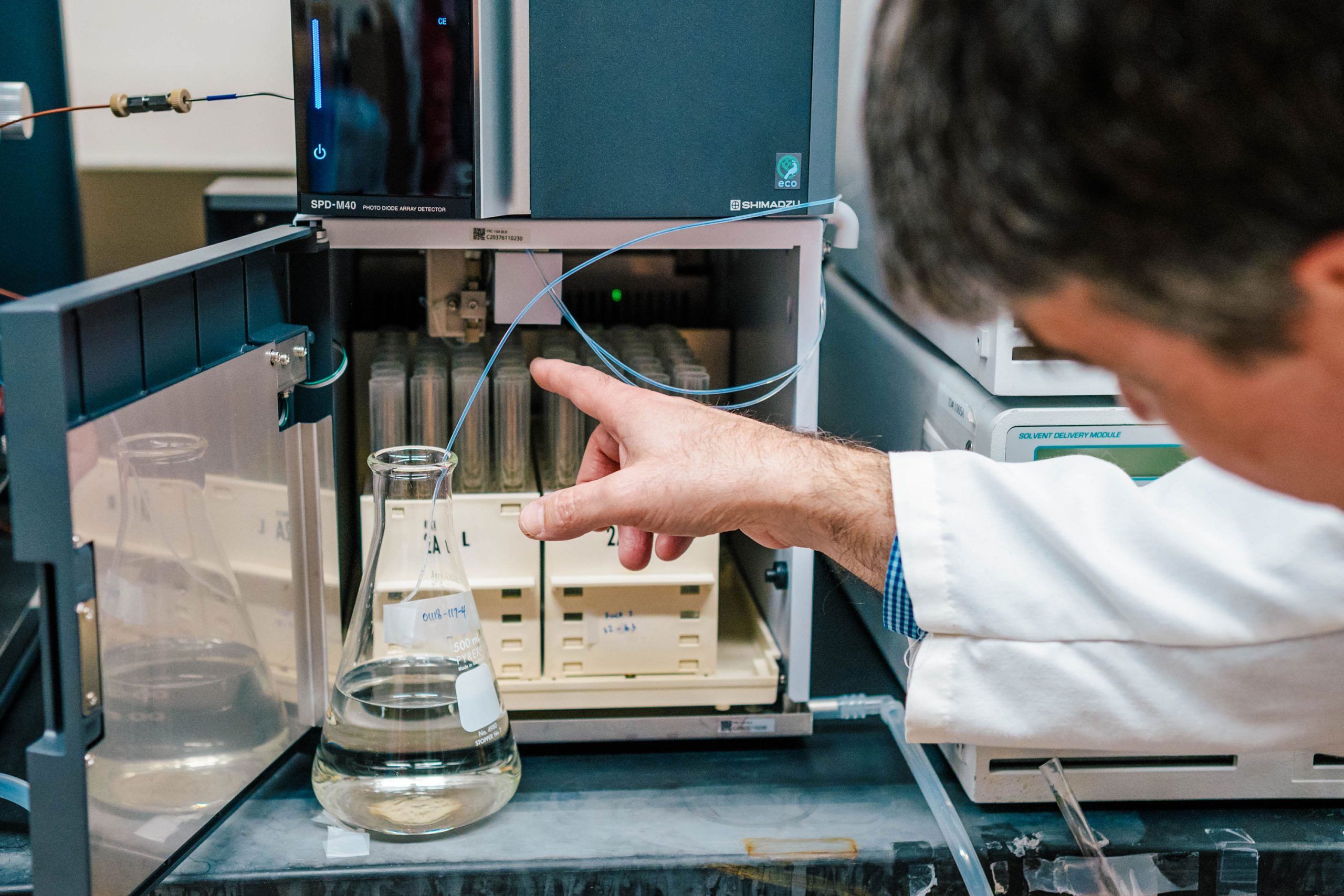 Laboratory equipment setup showing a person’s gloved hand pointing to a chromatography system with tubing connected to a glass flask containing clear liquid. The system includes labeled trays and a digital display.