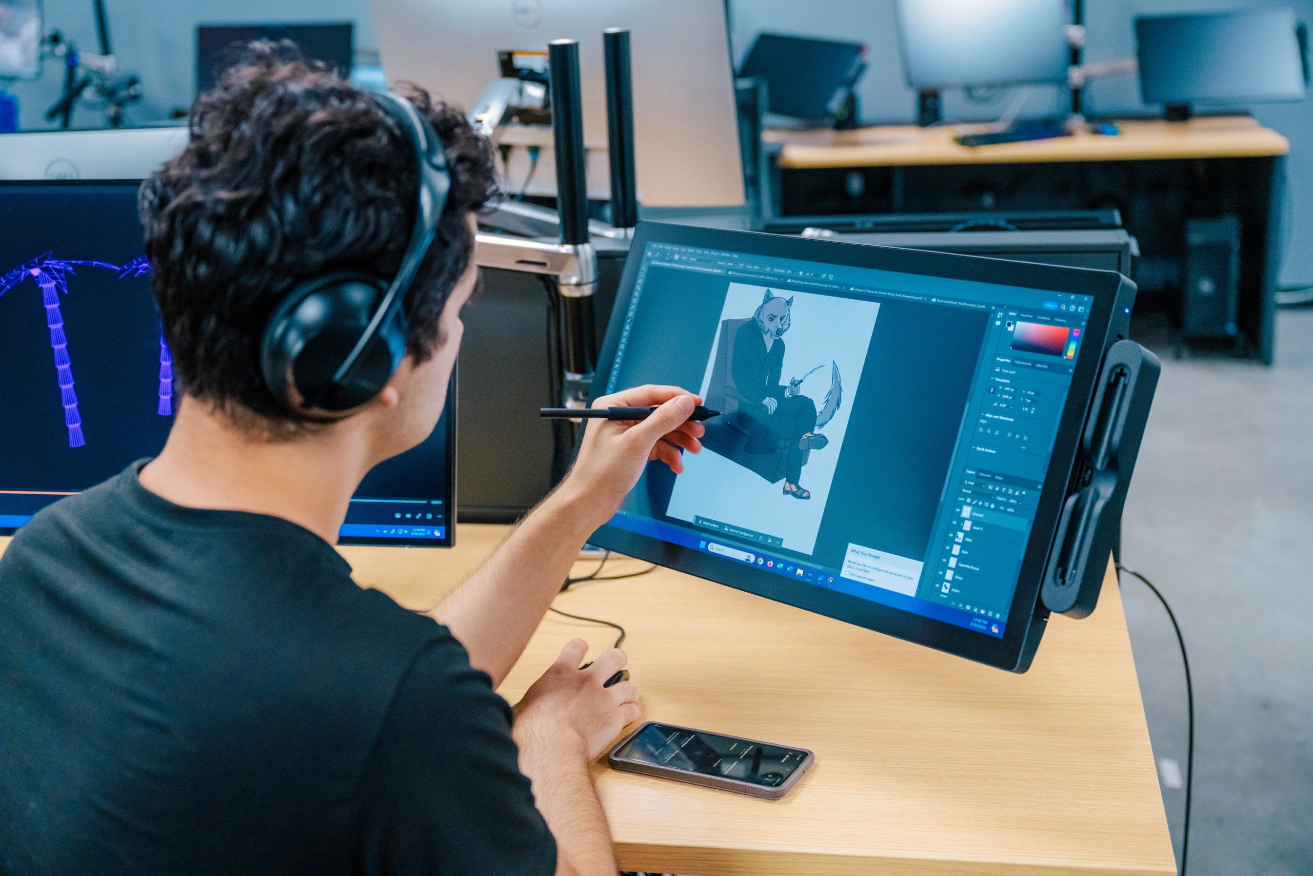 A person wearing headphones uses a stylus on a large drawing tablet to create digital artwork of a seated character in a design program. A smartphone rests on the desk, and multiple computer monitors are visible in the background, indicating a digital art or animation workspace.