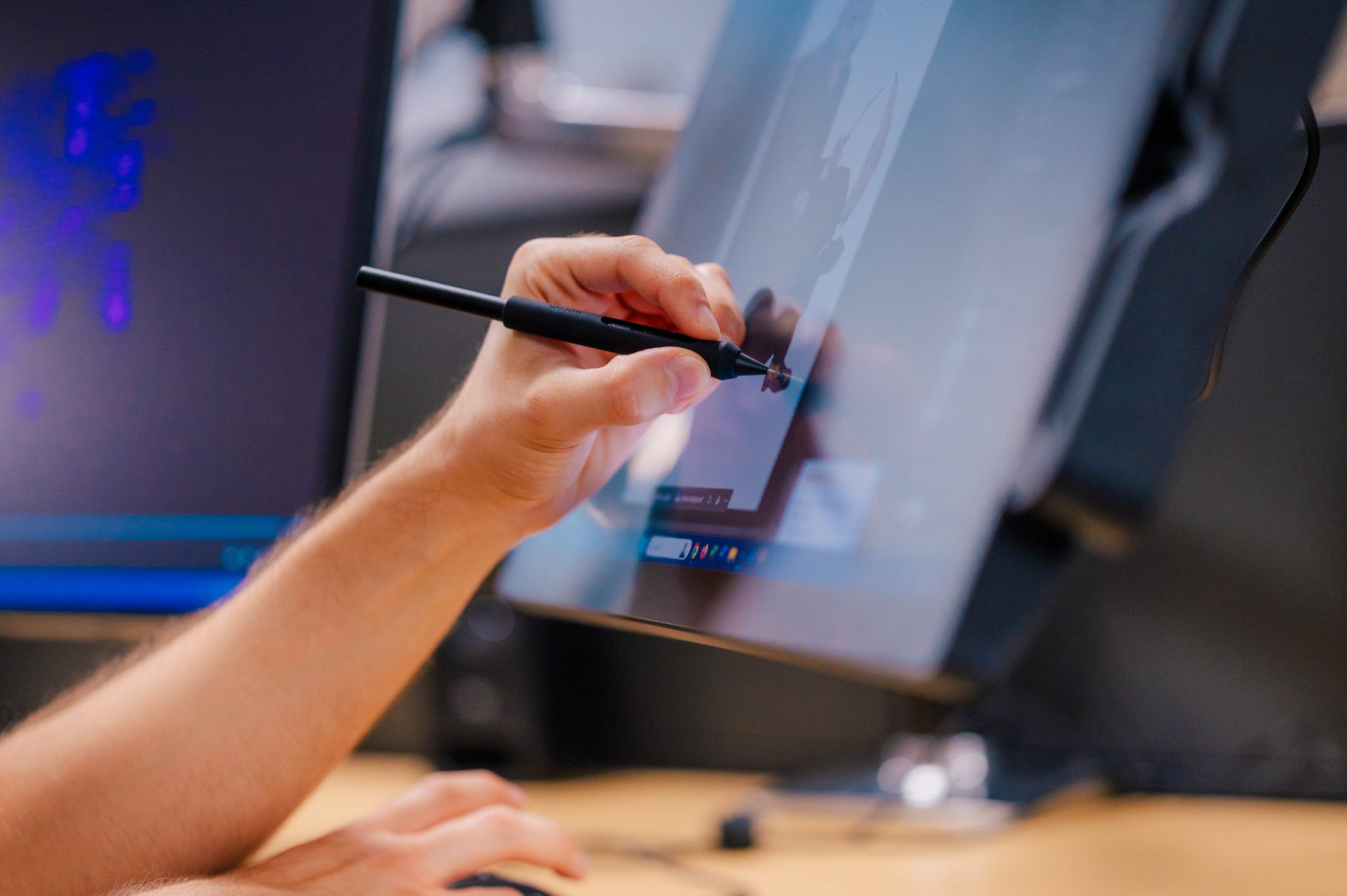 A close-up of a hand using a digital pen on a touchscreen monitor to create or edit artwork. The screen displays a grayscale image being drawn or refined, and the workspace includes another monitor and a keyboard in the background, suggesting a digital art or animation setup.