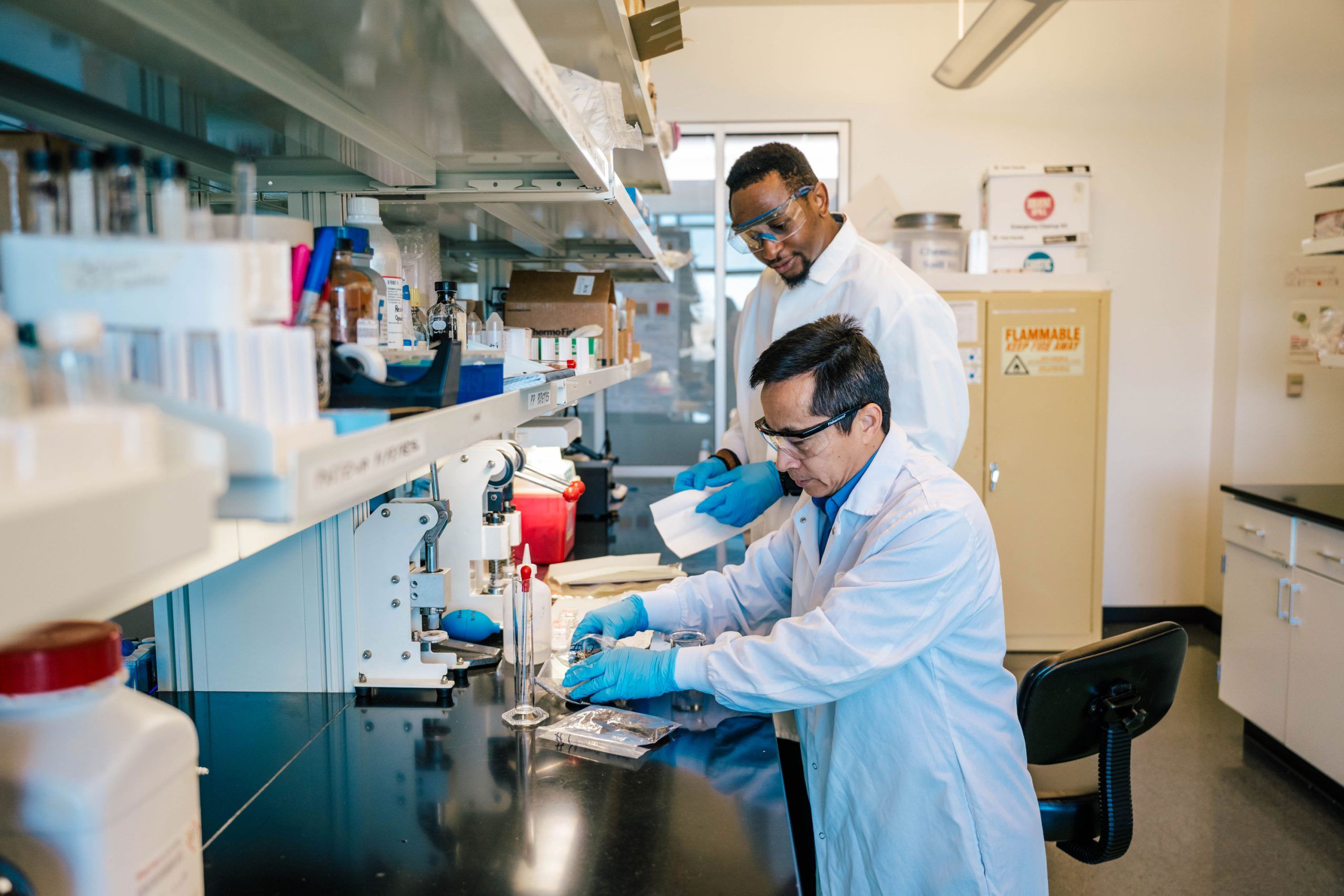Dr. Wei and his student in a laboratory wearing lab coats and blue gloves working at a black countertop. One is seated using a pipette with a tall glass cylinder, while the other stands nearby holding papers. Shelves above contain boxes and containers, and a cabinet labeled “Flammable Keep Fire Away” is visible in the background.