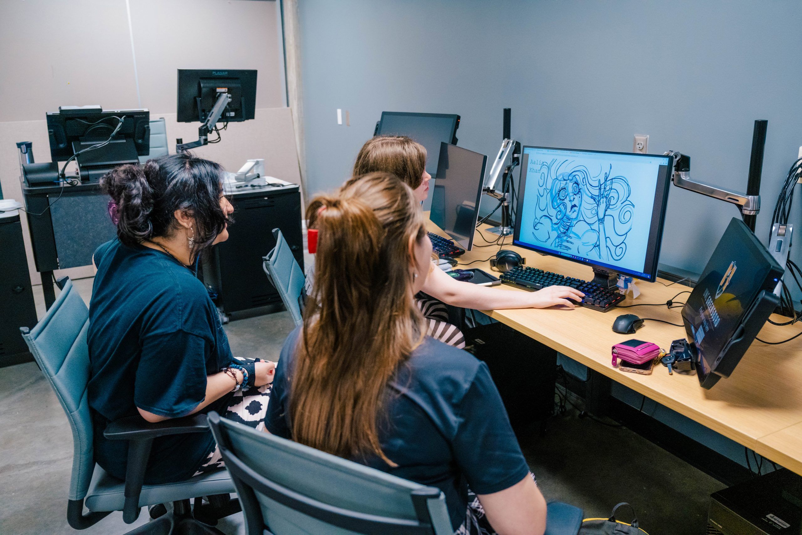 Three individuals sit at a workstation in a computer lab, looking at a large monitor displaying a digital illustration of a stylized character with flowing hair and intricate line art. The desk has multiple monitors, keyboards, and a small pink case. In the background, additional computer stations and equipment are visible, suggesting a collaborative digital art or animation environment.