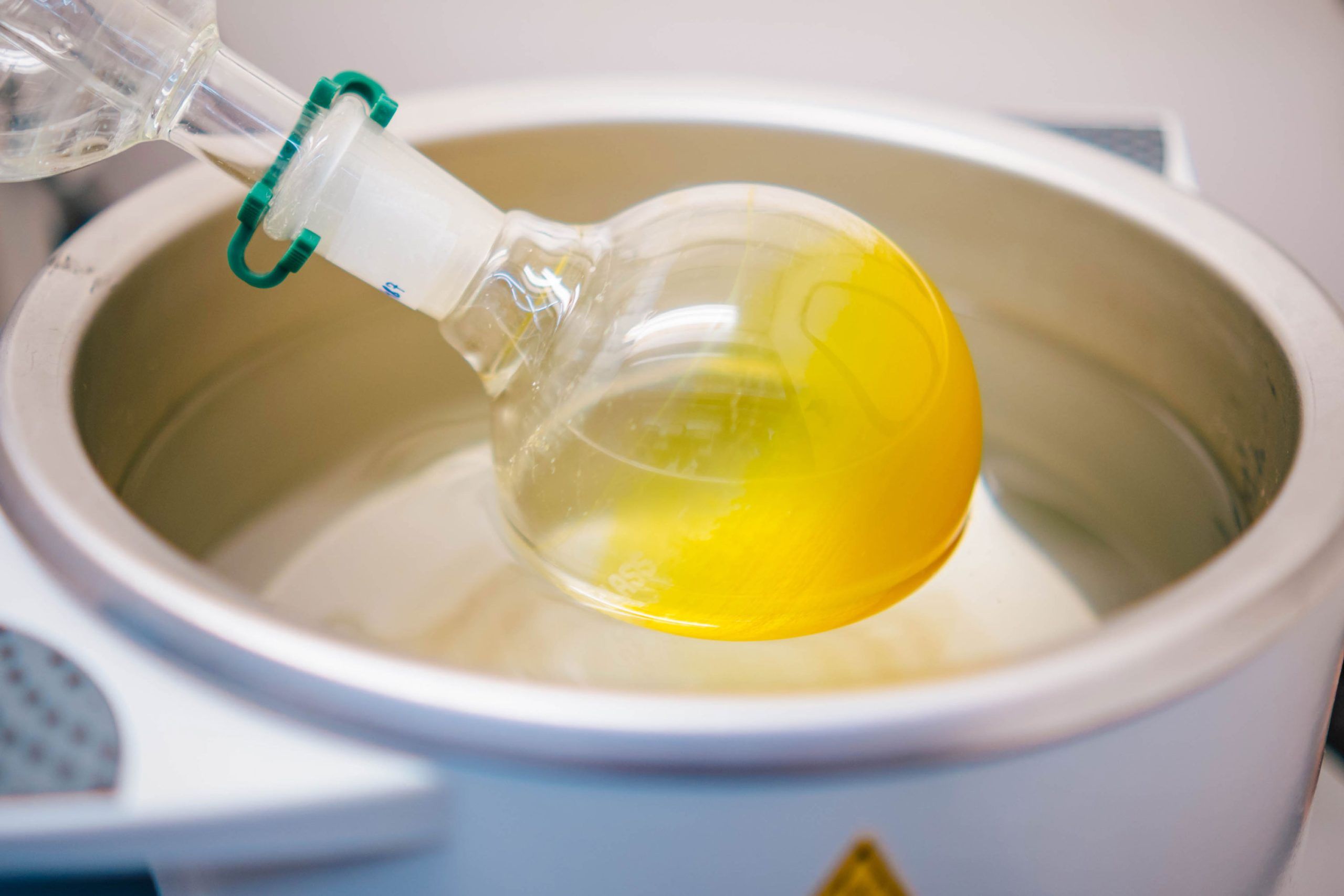 Close-up of a round-bottom glass flask containing bright yellow liquid, partially submerged in a metal water bath used for heating in a laboratory setting.