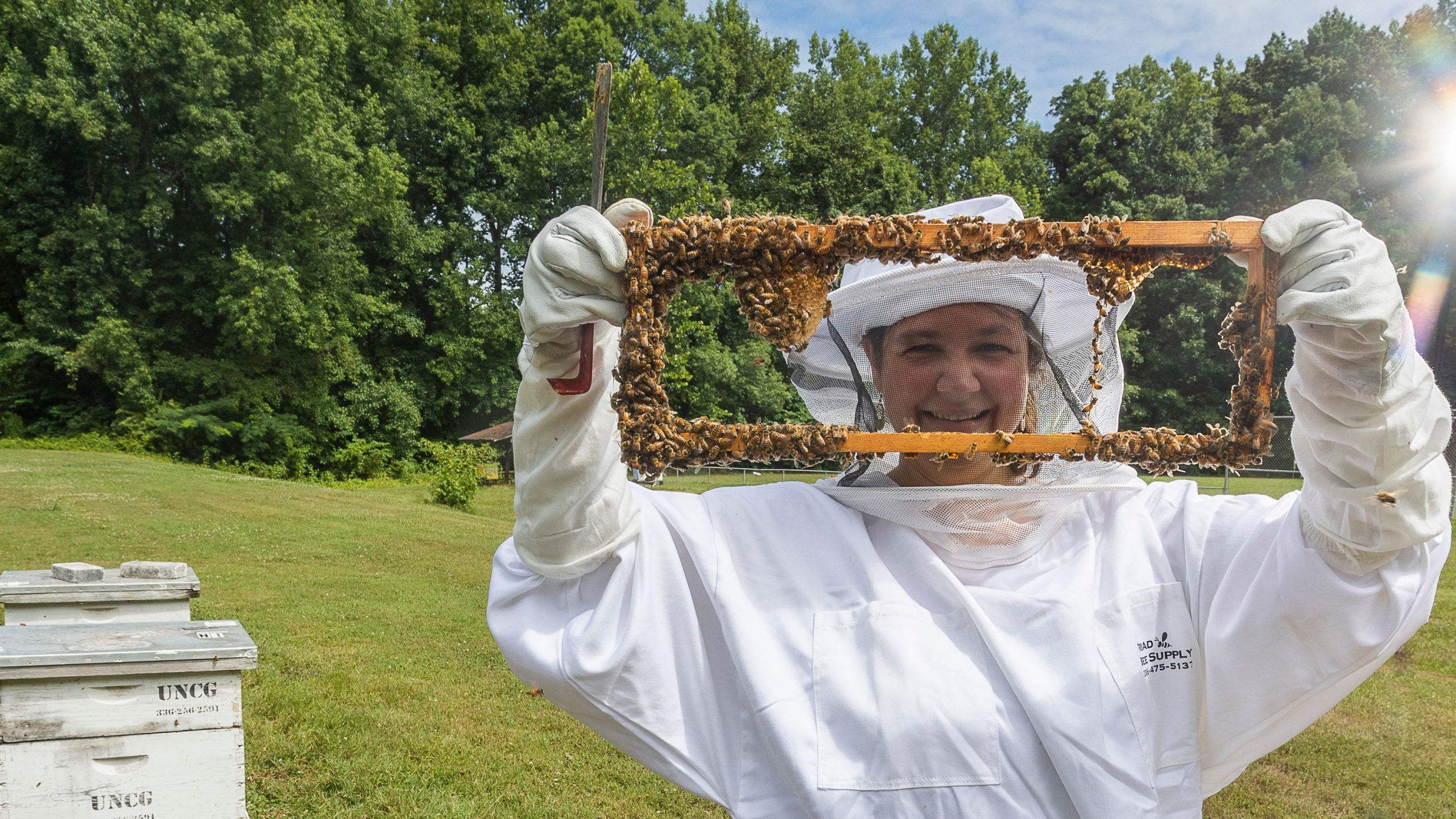 A person in beekeeping gear holding a rectangular object covered in bees