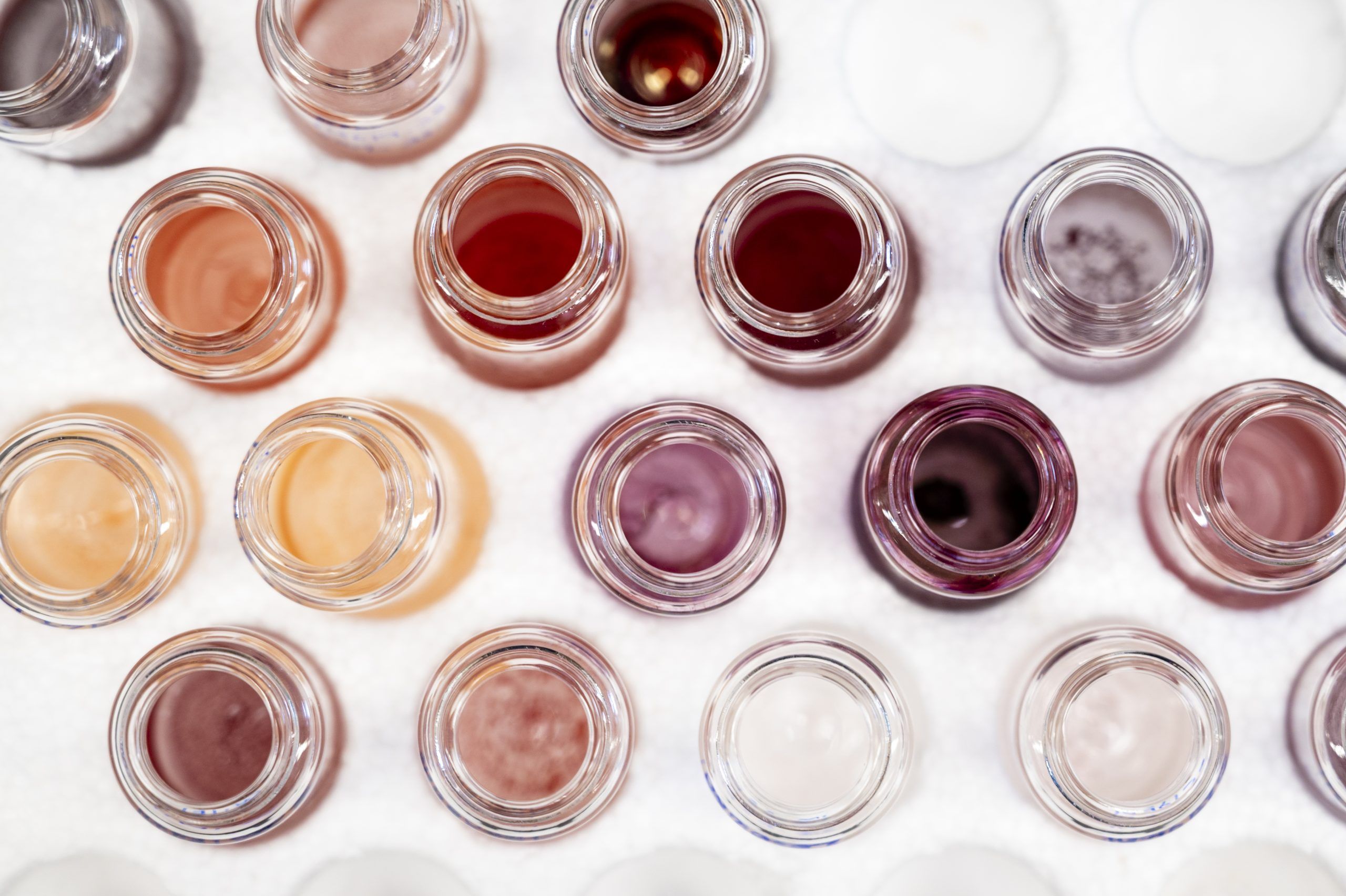 Top-down view of multiple small glass jars arranged in rows, each containing liquids in varying shades of beige, pink, and brown.