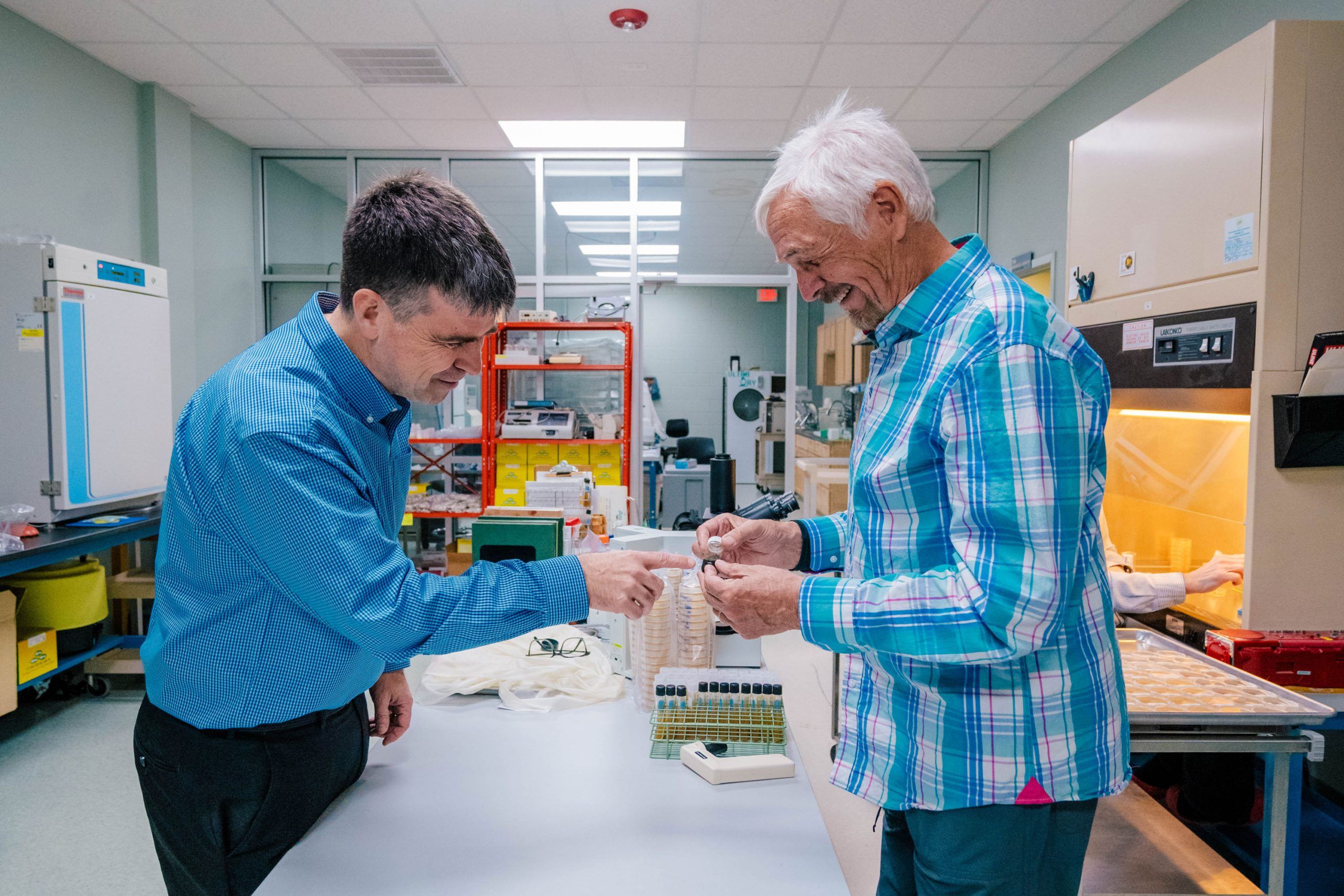 Dr. Oberlies and Cedric Pearce in a laboratory examining small sample containers on a workbench. The background includes lab equipment, shelving, and a fume hood with a yellow-lit workspace.
