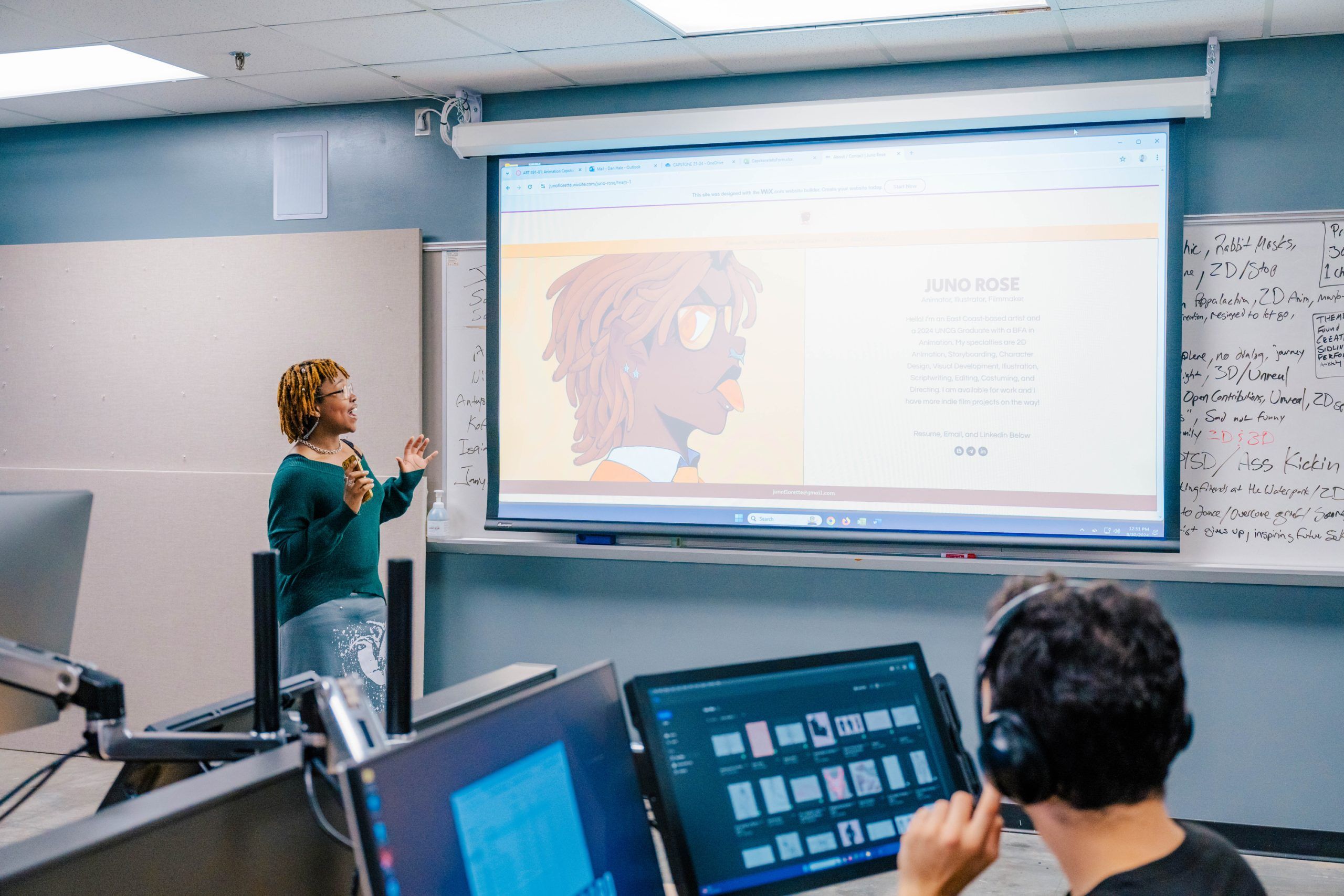 A classroom presentation where a person stands near a whiteboard and gestures toward a large projection screen displaying a digital illustration of a character labeled “JUNO ROSE” with descriptive text. In the foreground, computer monitors show design software, and another individual wearing headphones works at a desk. The setting is a modern computer lab with a collaborative learning environment.
