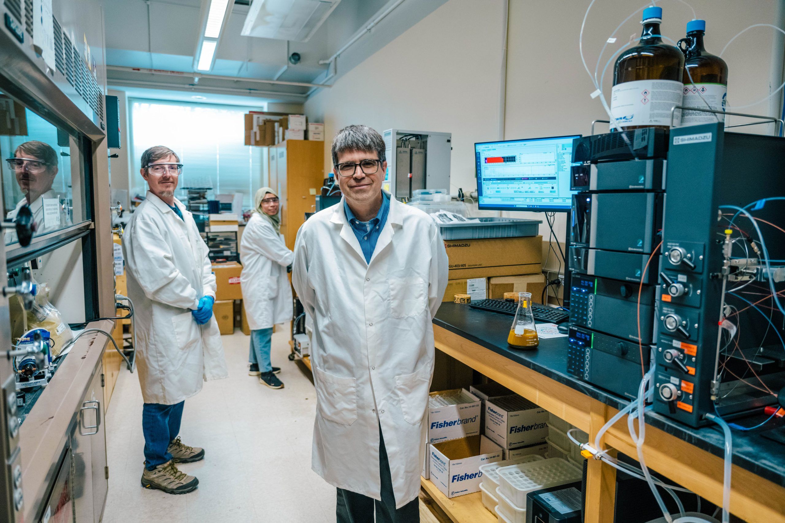 Laboratory with multiple pieces of analytical equipment, including a chromatography system connected to tubing and glass flasks. A computer monitor displays data charts, and shelves hold large reagent bottles. Cardboard boxes labeled “Fisherbrand” are stacked under the counter.