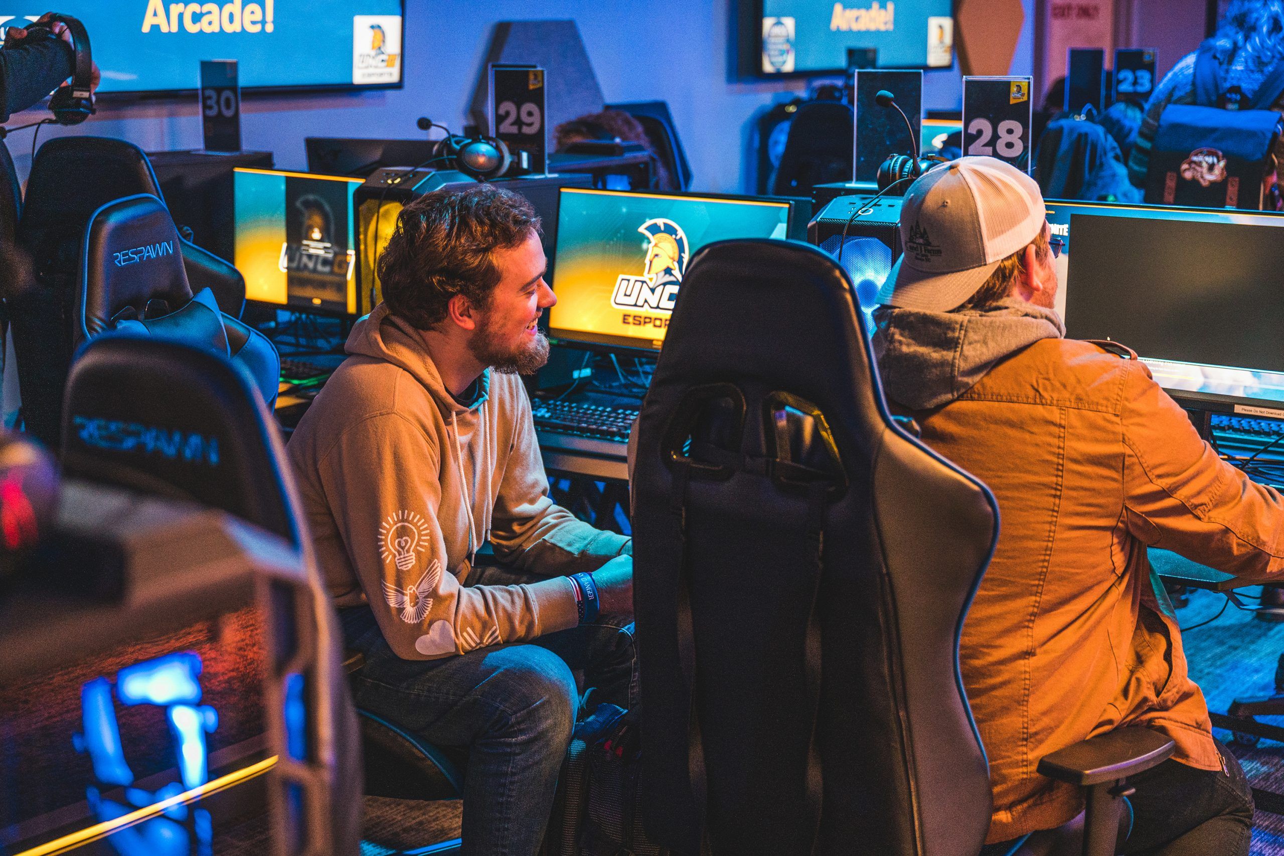 Two individuals seated at gaming stations in an esports arena, wearing casual clothing and using monitors, keyboards, and headsets, with UNCG Esports branding visible on screens and walls.