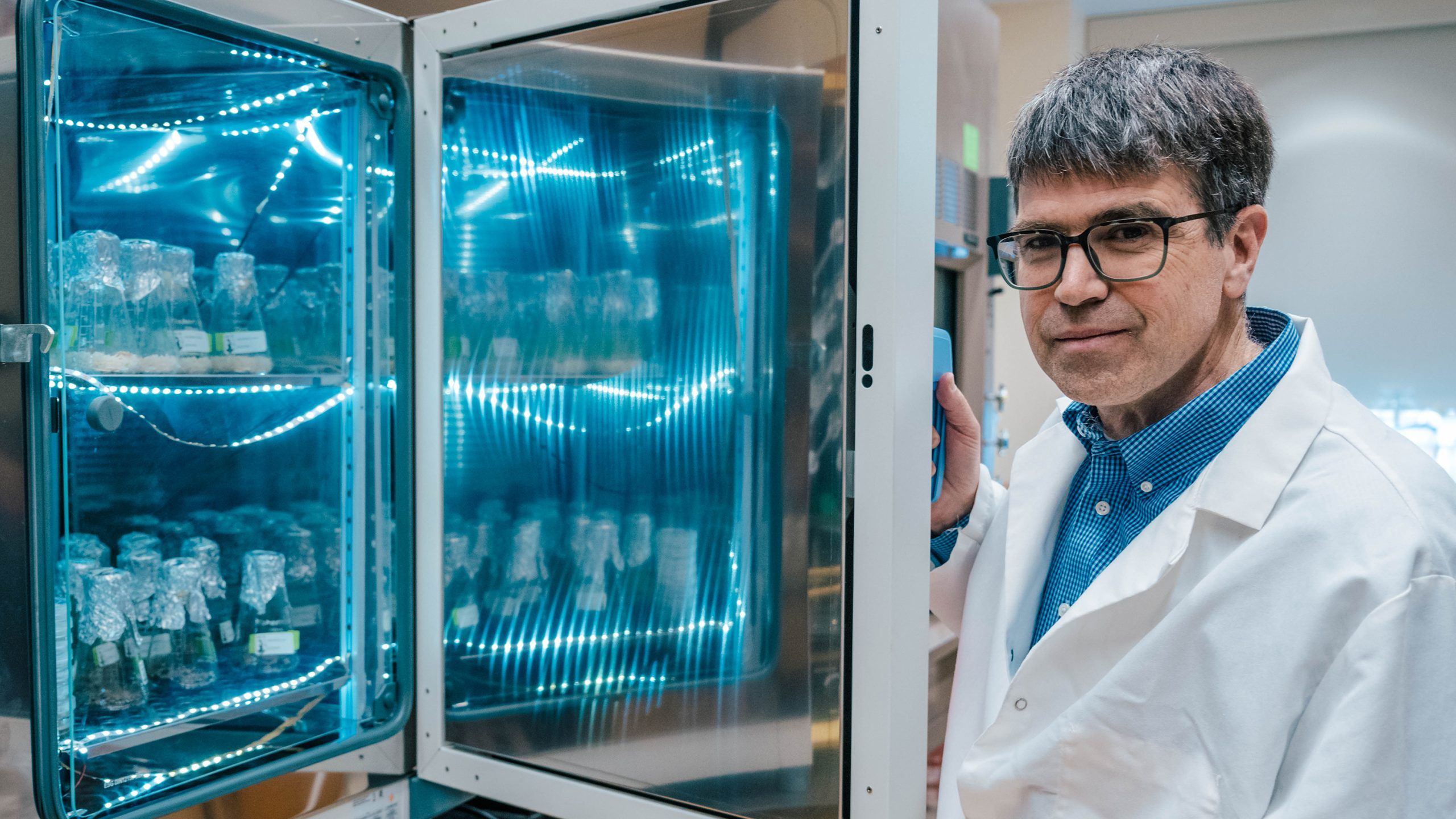 Dr. Oberlies in a lab coat standing next to an open refrigerated storage unit containing multiple glass bottles with foil covers, illuminated by blue LED lights.