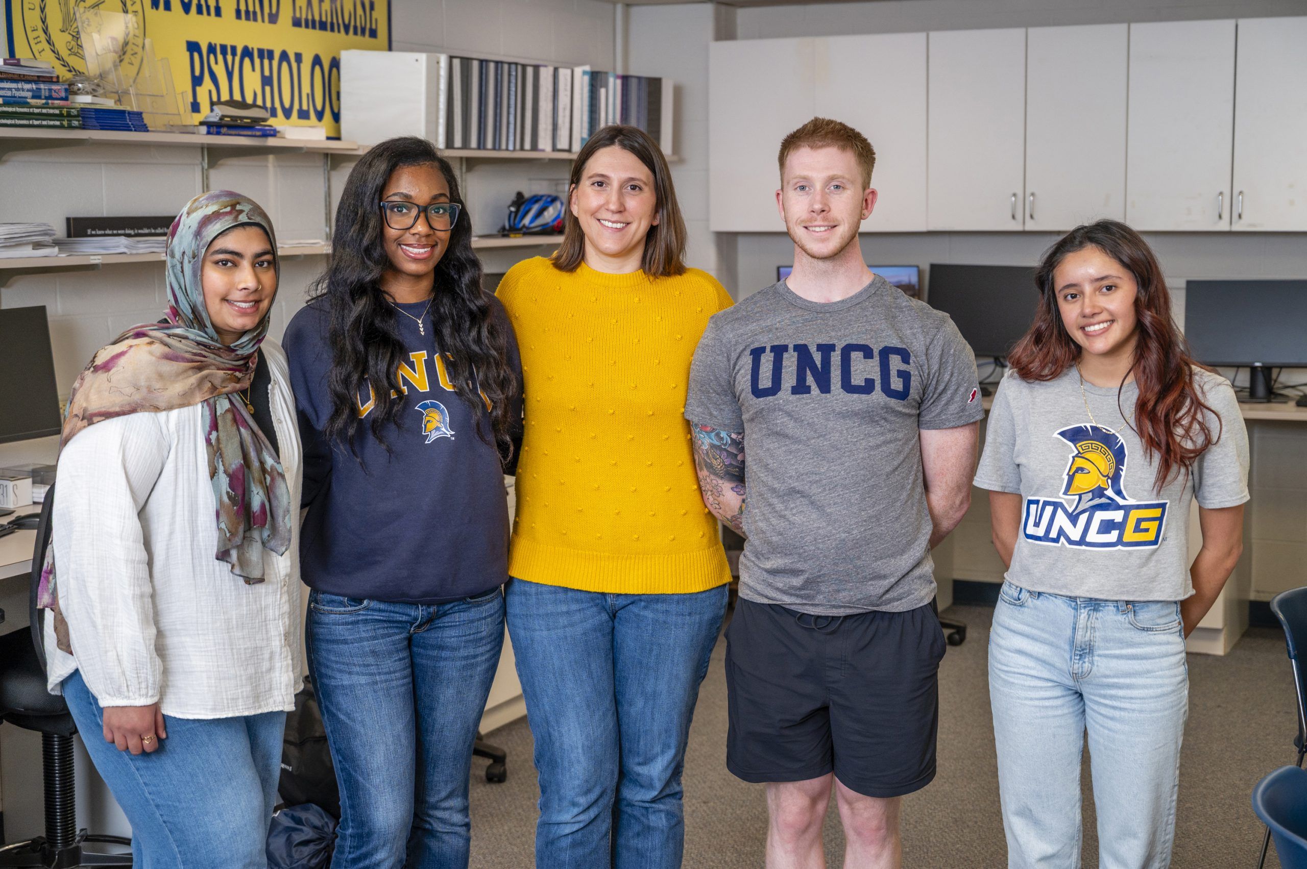 Five people stand side by side in a classroom or office setting, posing for a group photo. They are wearing casual clothing, including UNCG-branded shirts and a bright yellow sweater. The background shows desks with computer monitors, shelves with binders, and a banner partially visible that reads “Psycholog.”