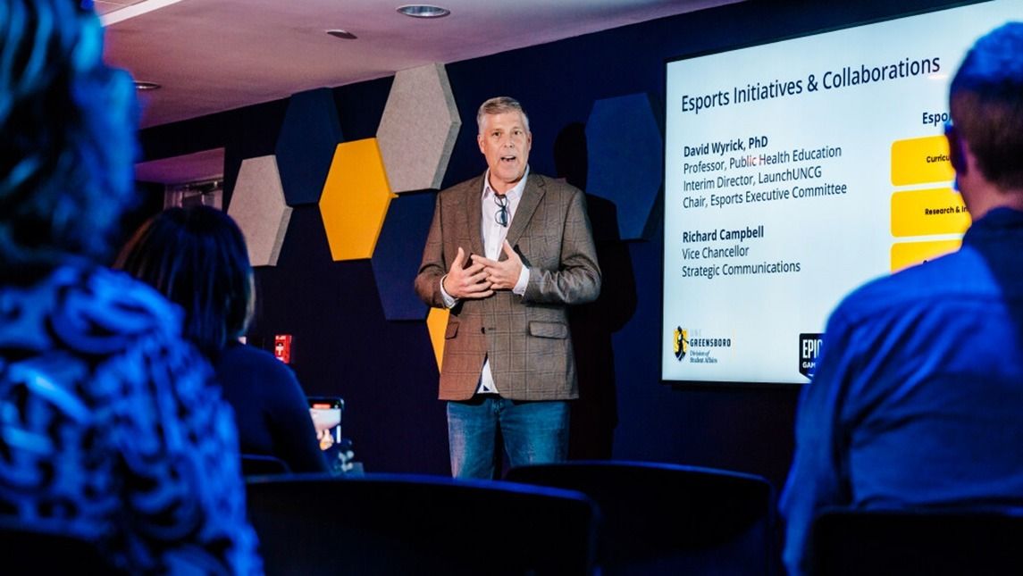 David Wyrick giving a presentation in a room with hexagonal wall panels, standing near a large screen displaying a slide titled ‘Esports Initiatives & Collaborations’ with names and roles listed.